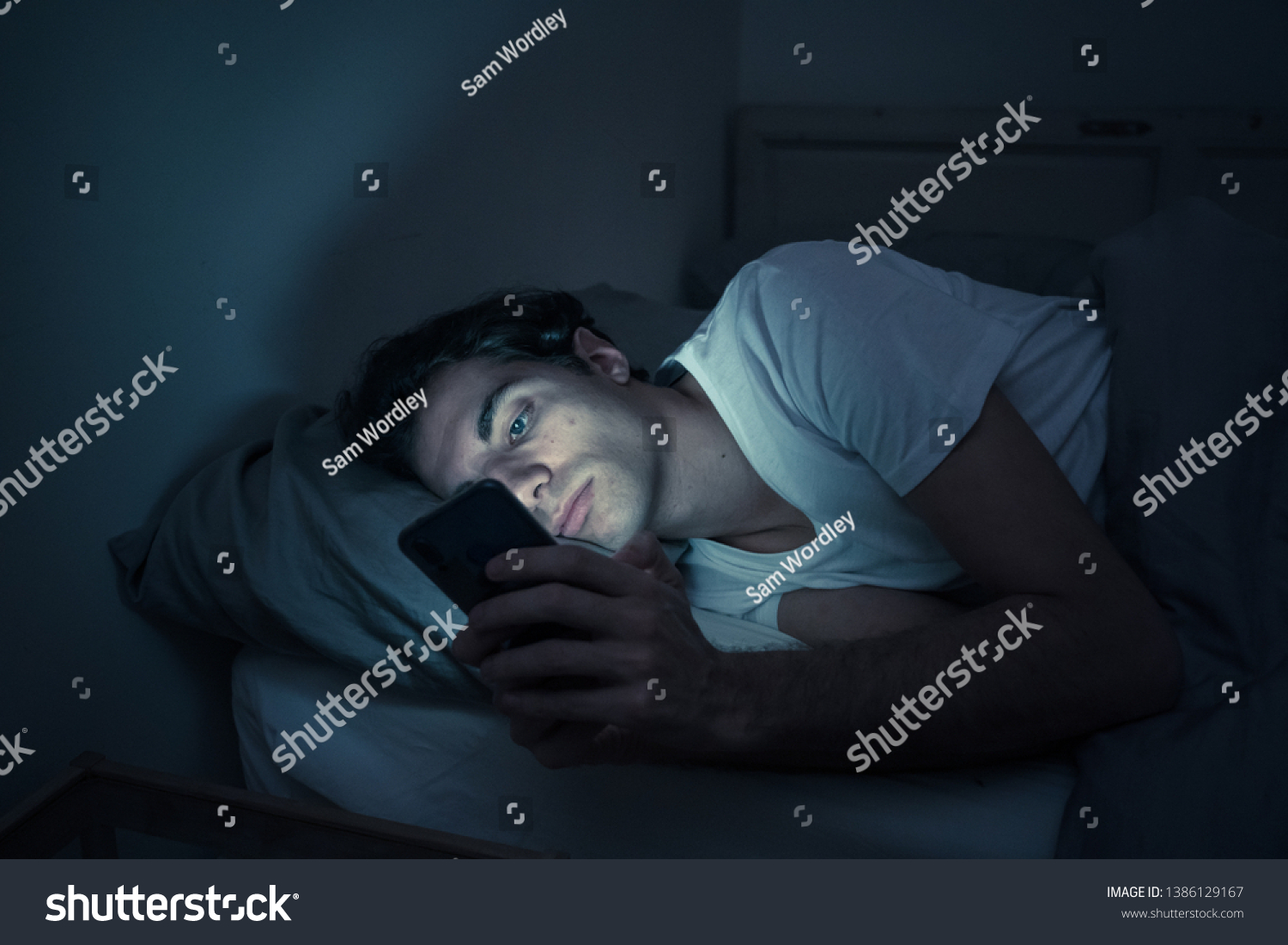 Addicted man chatting and surfing on the Internet with smart phone late at night in bed. Bored, sleepless and tired in dark room with moody light. In insomnia and mobile addiction concept. #1386129167