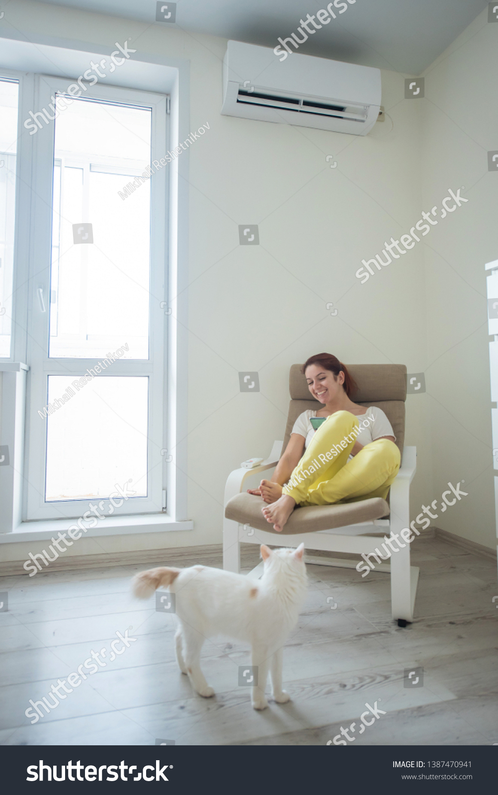 Red-haired girl in bright clothes and barefoot sits in a chair with a phone in her hands under the air conditioner. Young woman sitting in an armchair with a cat #1387470941
