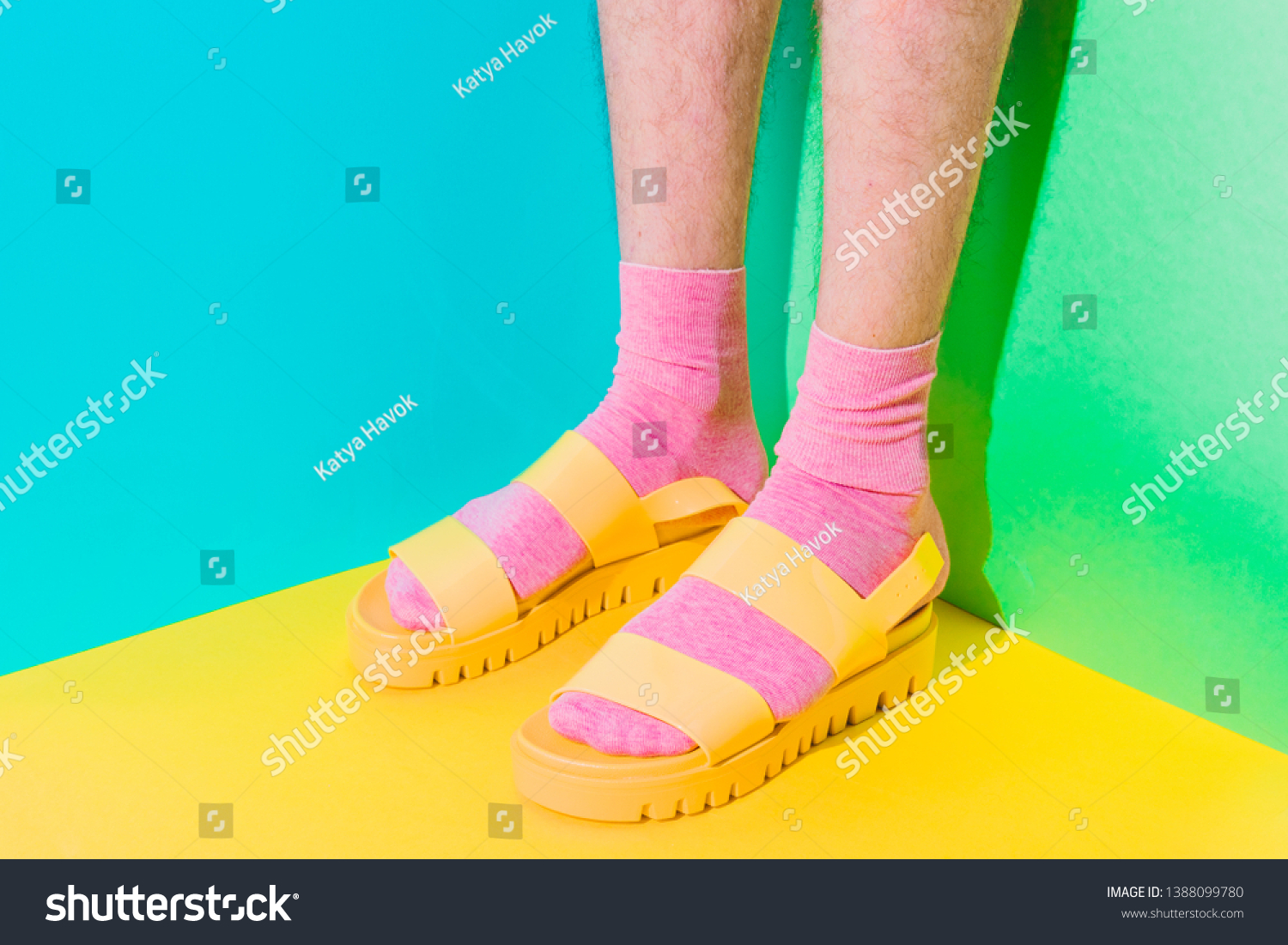 Male hairy legs in socks staying in women's sandals on bold background in the corner with strong shadows. Minimal pride concept. Body part #1388099780