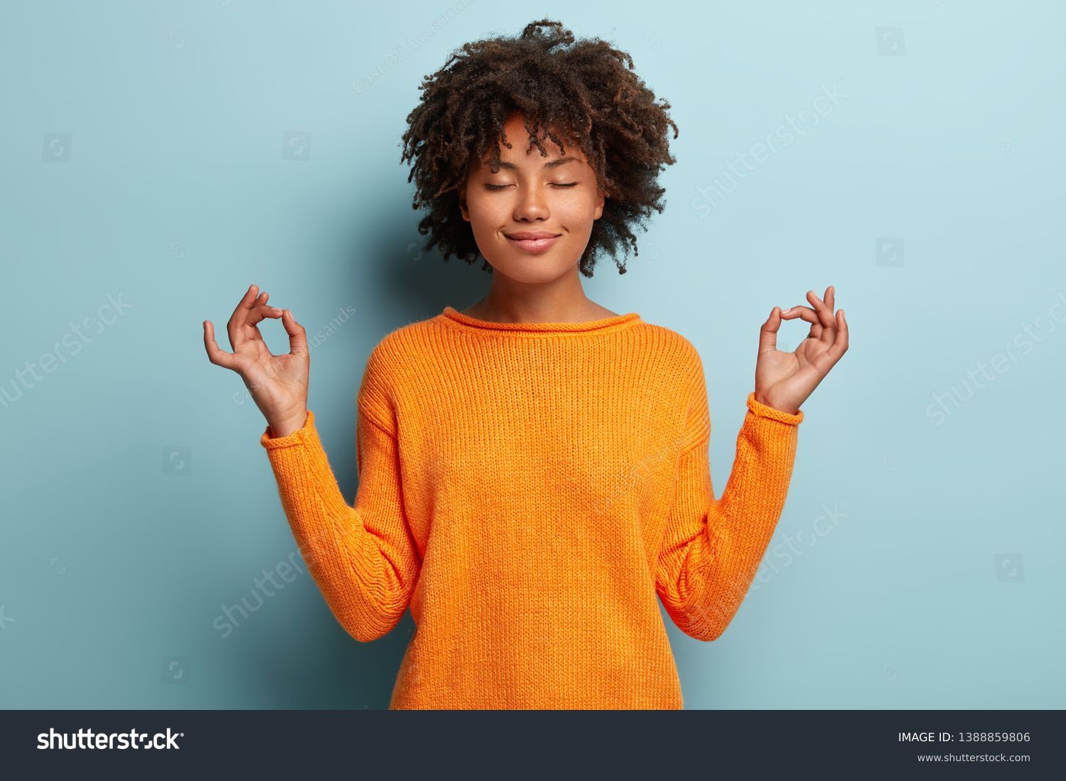 Mindful peaceful Afro American woman meditates indoor, keeps hands in mudra gesture, has eyes closed, tries to relax after long hours of working, holds fingers in yoga sign, isolated on blue wall #1388859806