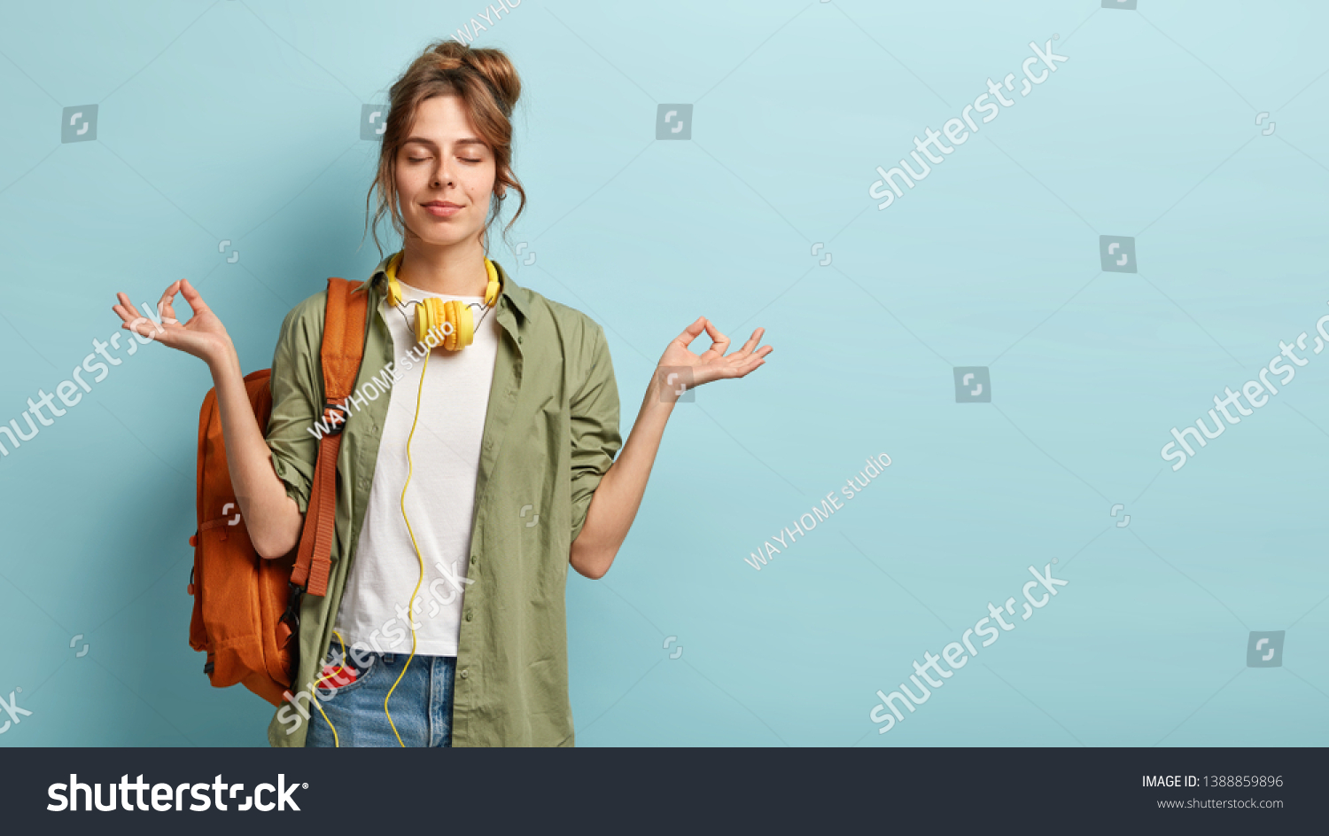 Photo of relaxed student keeps hands in mudra gesture, keeps eyes closed, listens peaceful music, headphones on neck, wears shirt and jeans, has rucksack, isolated on blue background with free space #1388859896