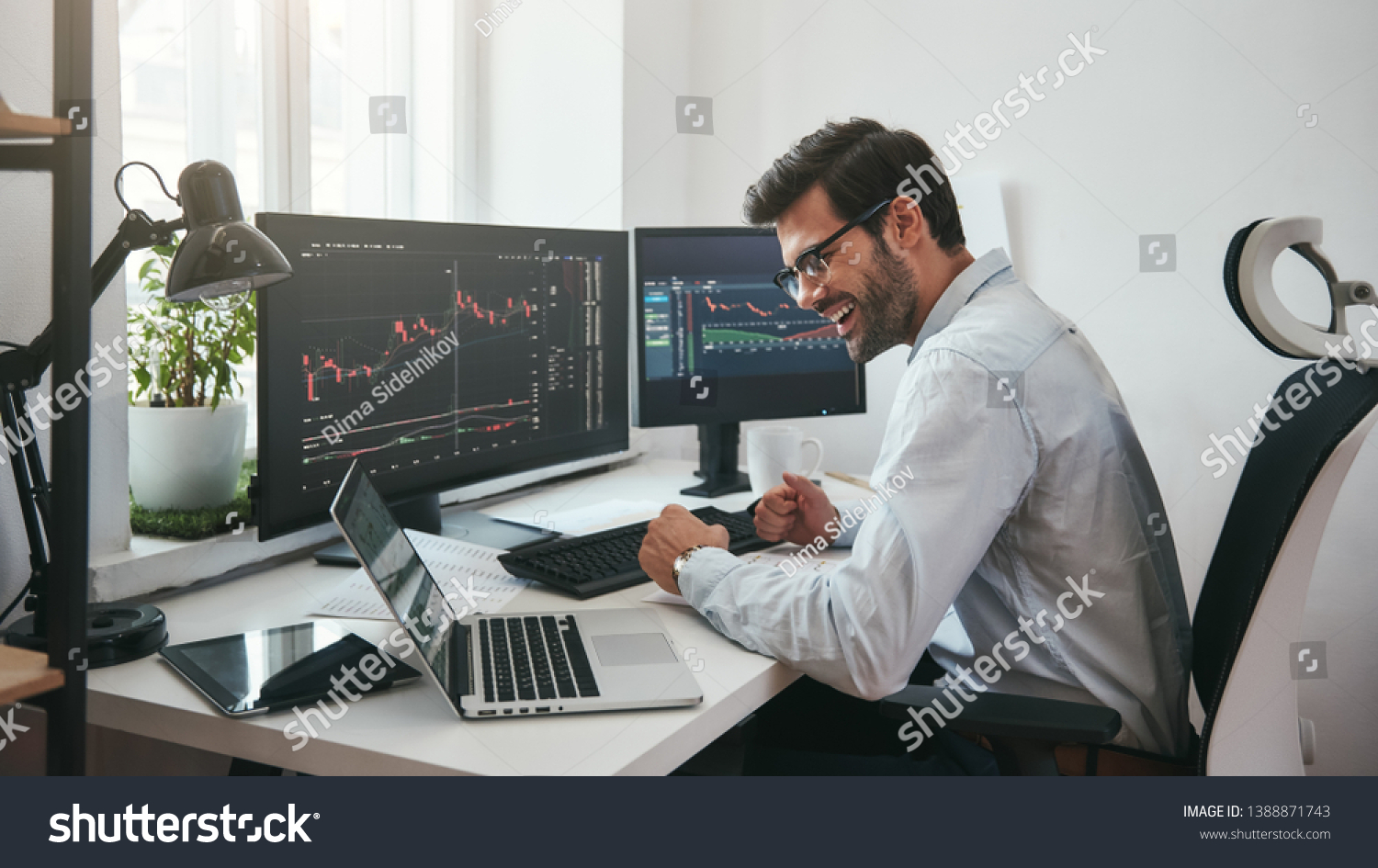 Lucky day. Happy young businessman or trader in formalwear and eyeglasses using laptop and smiling while sitting in his modern office #1388871743