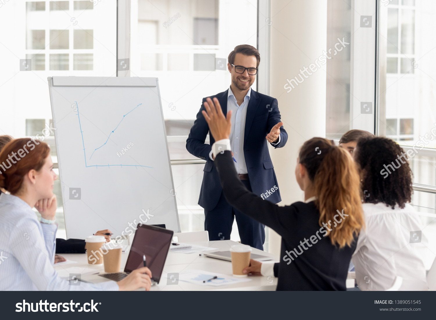 Smiling millennial male coach or presenter make flip chart presentation ask question during work training, motivated confident female employee raise hand answer engaged in teamwork at seminar #1389051545