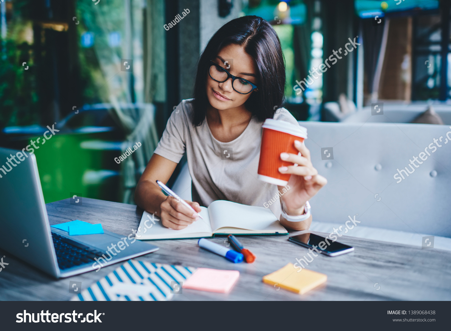 Concentrated female student writing in notebook while learning with cardboard coffee cup in cafe, pensive woman freelancer noting information for planning project doing remote job via laptop computer #1389068438