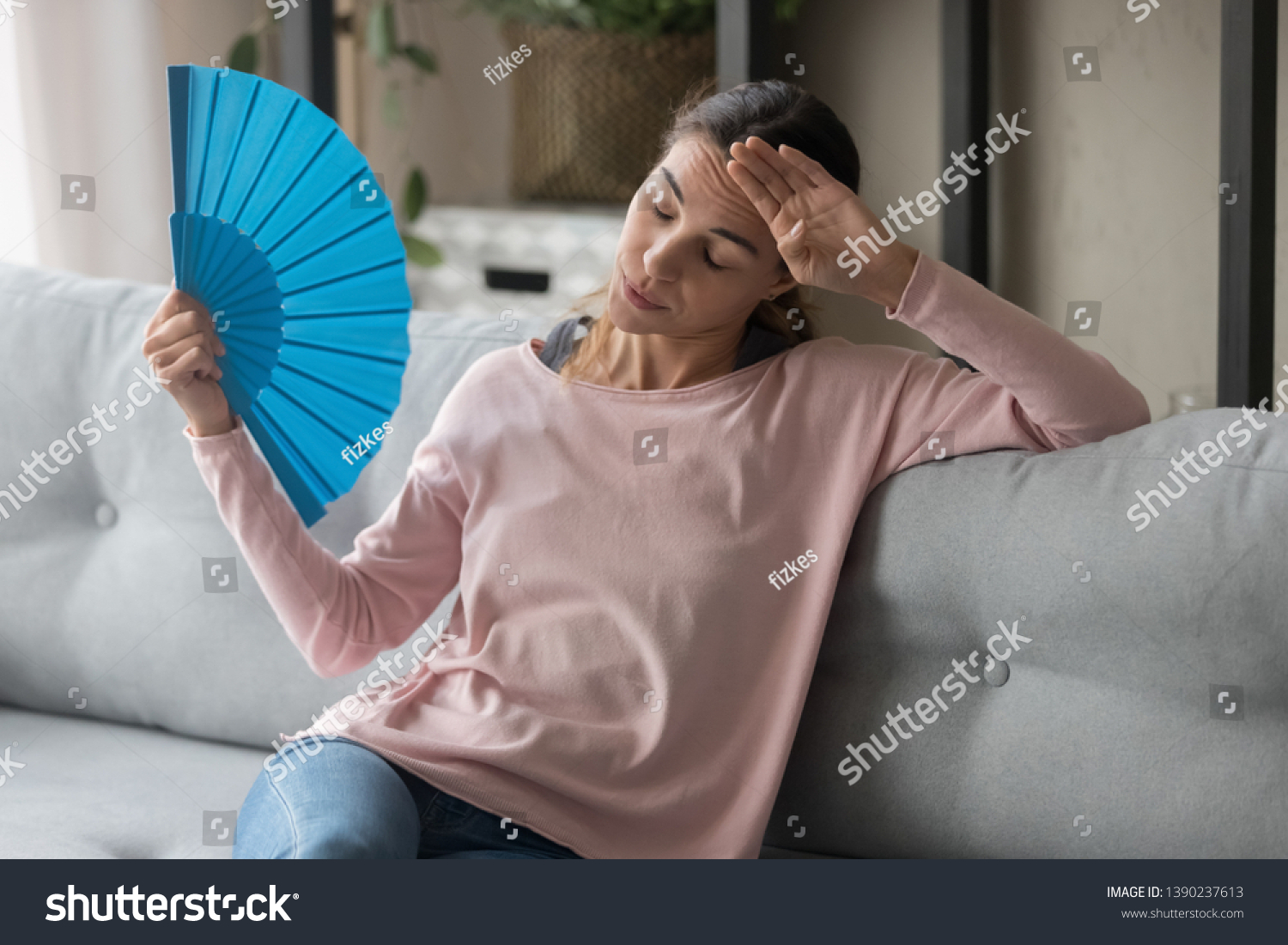 Overheated female sitting on couch in living room at hot summer weather day feeling discomfort suffers from heat waving blue fan to cool herself, girl sweating dwelling without air conditioner concept #1390237613