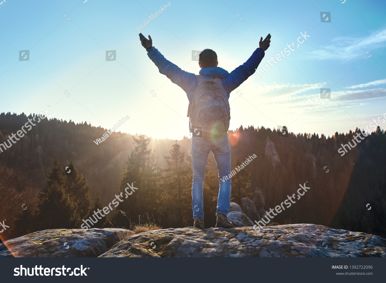 Male traveler standing on the cliff against wooded hills and cloudy sky at sunrise. Man stands hands up, greeting the sun. Adult tourist with backpack looking to misty hilly valley below. Location #1392722096