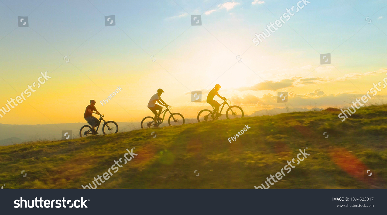 AERIAL LENS FLARE COPY SPACE SILHOUETTE: Fit tourists riding electric bicycles along a grassy path on a beautiful sunny spring day. Cinematic shot of three friends enjoying a scenic mountain bike ride #1394523017