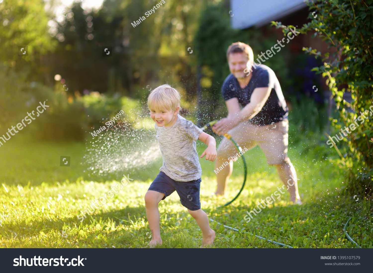 Funny little boy with his father playing with garden hose in sunny backyard. Preschooler child having fun with spray of water. Summer outdoors activity for kids. #1395107579