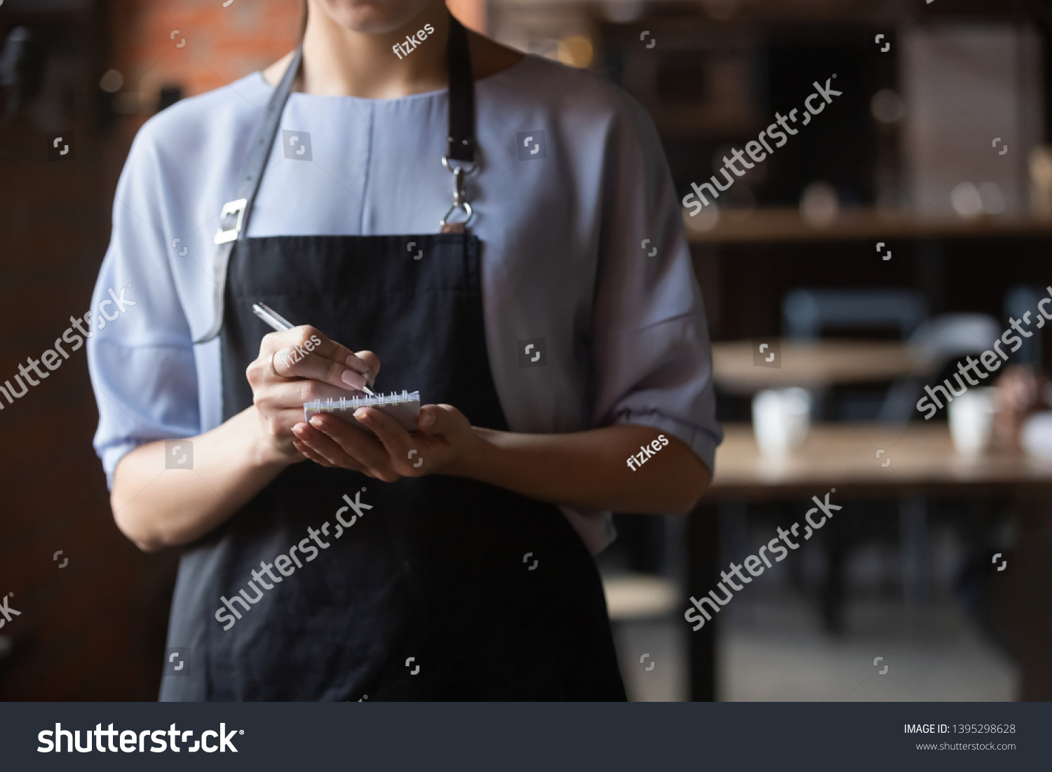Close up attractive waitress wearing black apron standing in cozy coffeehouse, female cafe worker with notebook in hands waiting for, ready to take customers, guests order in restaurant #1395298628