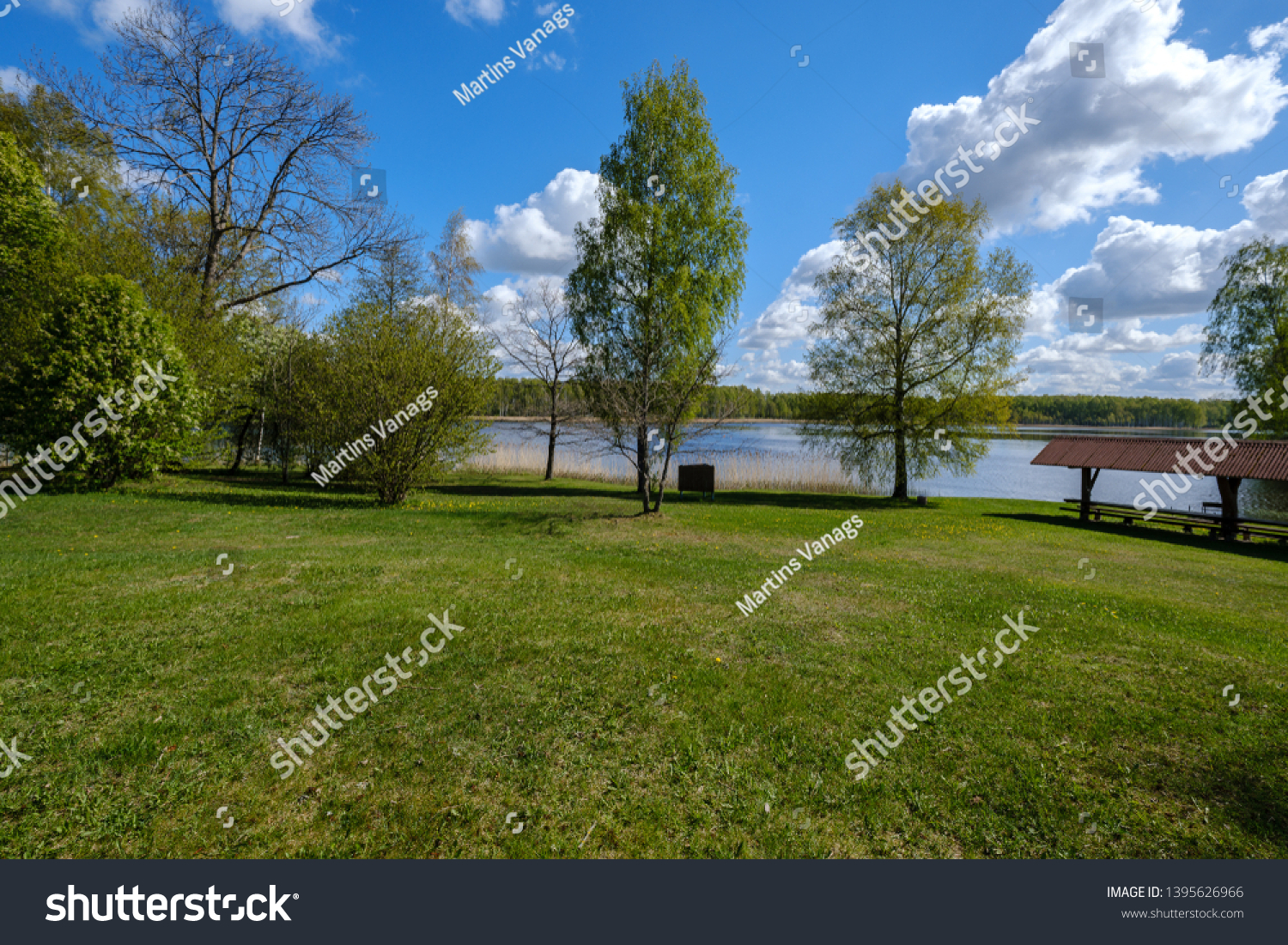 recreation camping area by the blue lake in sunny summer day on the shore of water body with trees, green meadow with dandelions and boats on the shore #1395626966