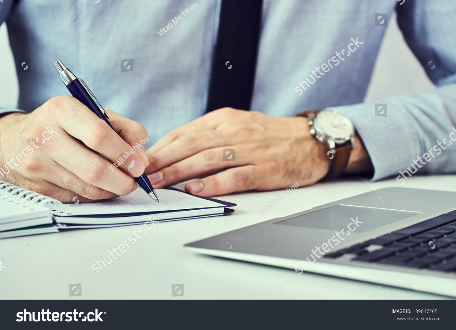 Businessman hand writing note on a notebook. Business man working at office desk. Close up of empty notebook on a blackboard with office supplies. #1396472651