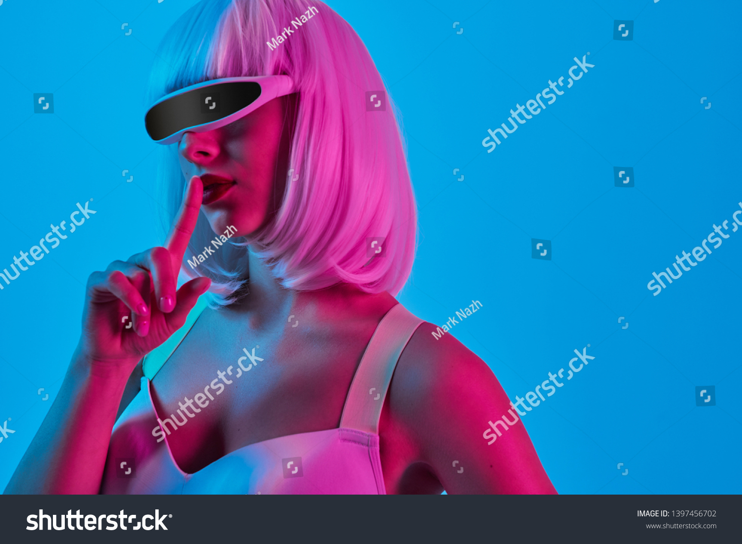 Futuristic alien woman in digital goggles keeping finger near lips and asking to be silent while standing under neon light on blue background #1397456702