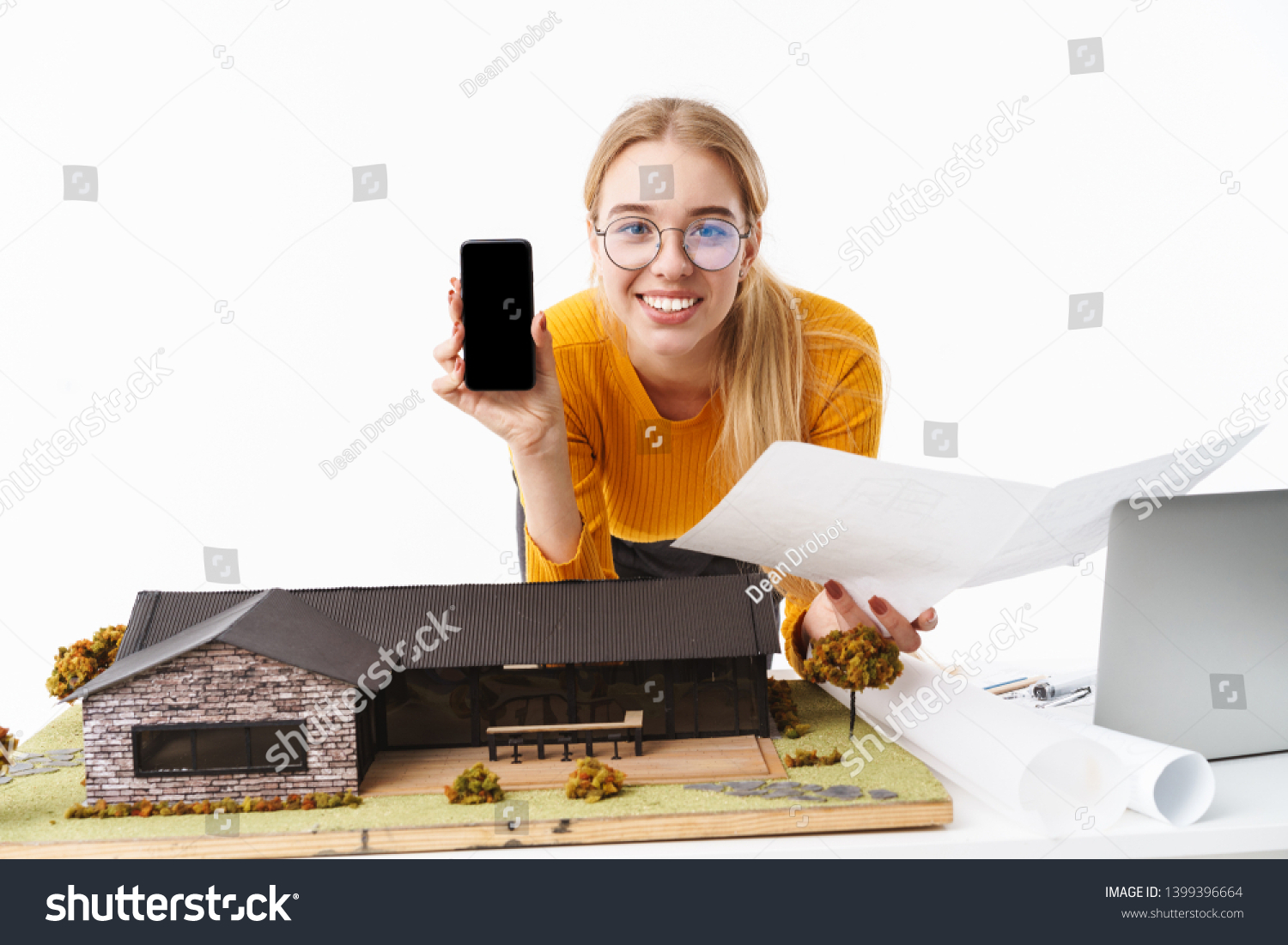 Beautiful young blonde woman architect standing isolated over white background, working on a new project at the desk with laptop, showing blank screen mobile phone #1399396664