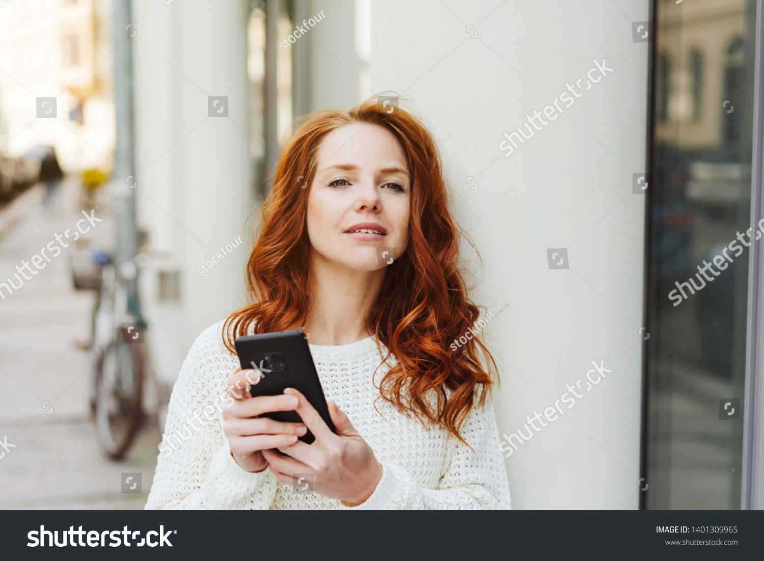 Young woman waiting in town for a meeting holding her mobile phone and peering expectantly down the street #1401309965
