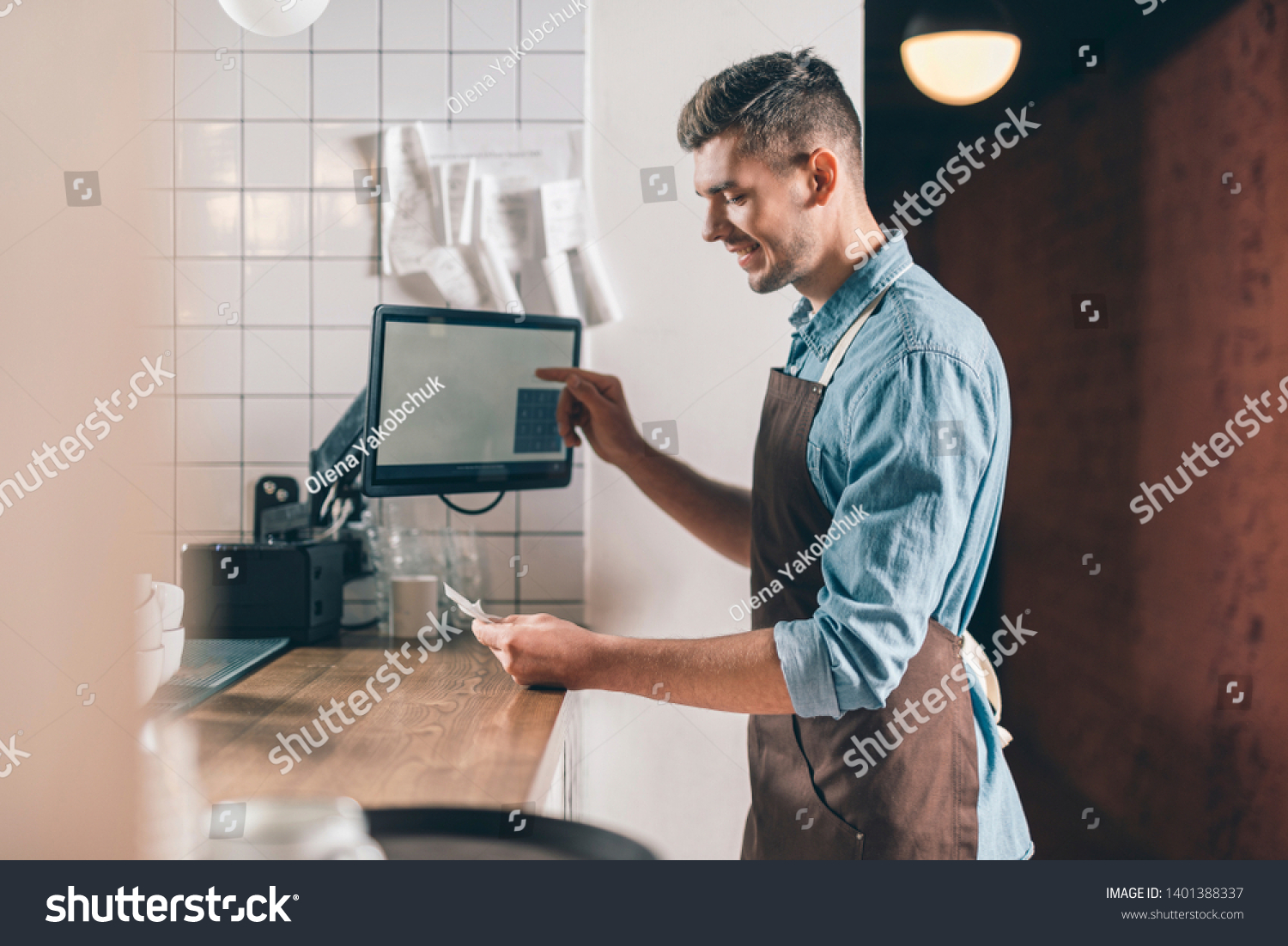 Checking the bill. Positive young waiter in a brown apron smiling and looking at the bill while standing at the bar counter #1401388337