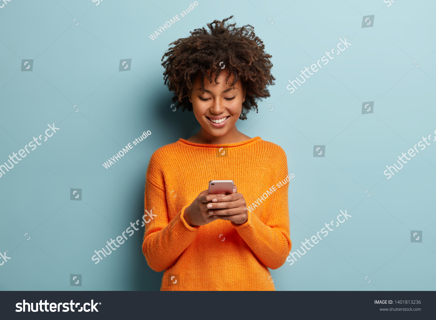 Satisfied hipster girl with Afro haircut, types text message on cell phone, enjoys online communication, types feedback, wears orange jumper, isolated on blue studio wall. Technology concept #1401813236