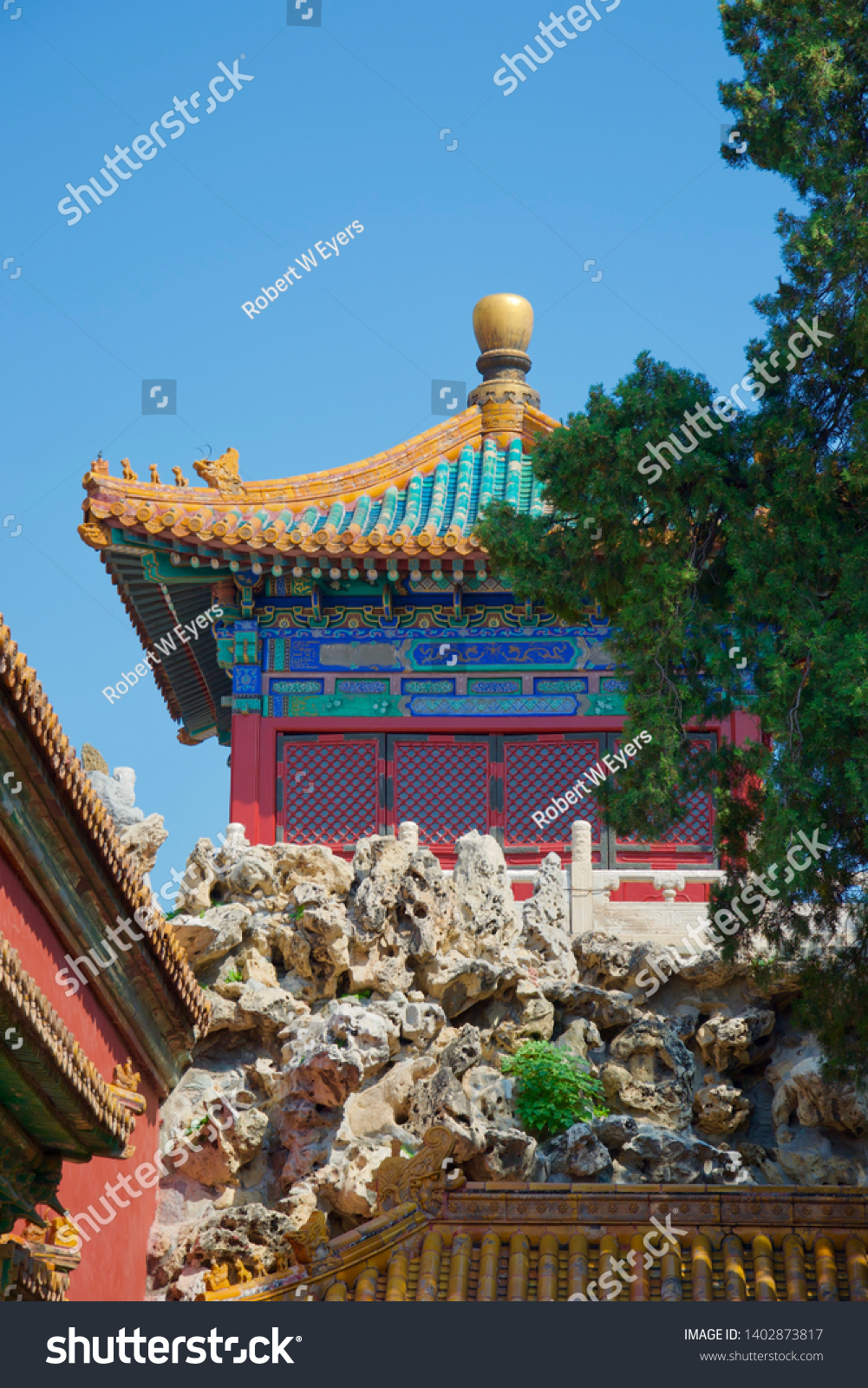A sunlit rockery and tree in foreground, with a vibrant, brightly painted Chinese temple (pagoda) in gold, blue, green and red against bright sky #1402873817
