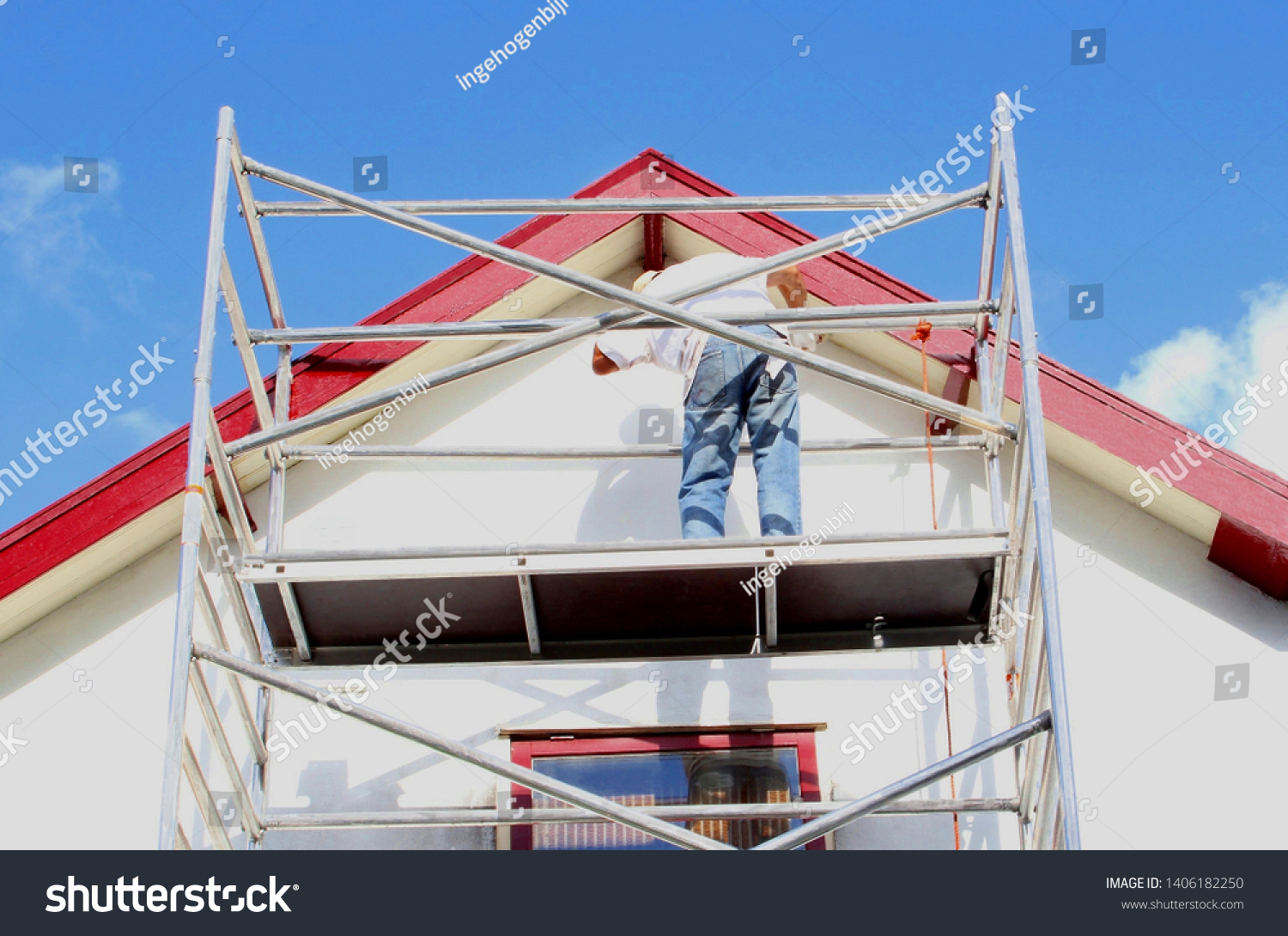 Professional workman is painting exterior walls and wooden window frames of ancient house at scaffold tower, outside home renovation in close up under sunny blue sky #1406182250