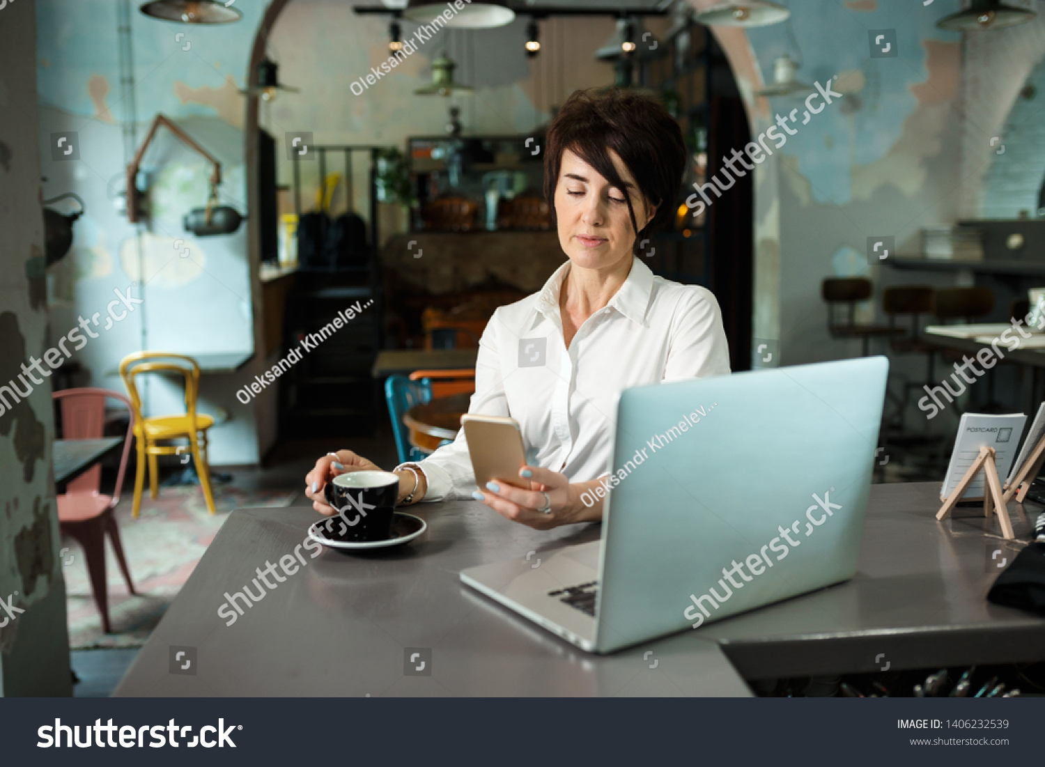 A middle-aged woman sits in a cafe, drinks coffee and works at a computer. Woman holding a phone #1406232539