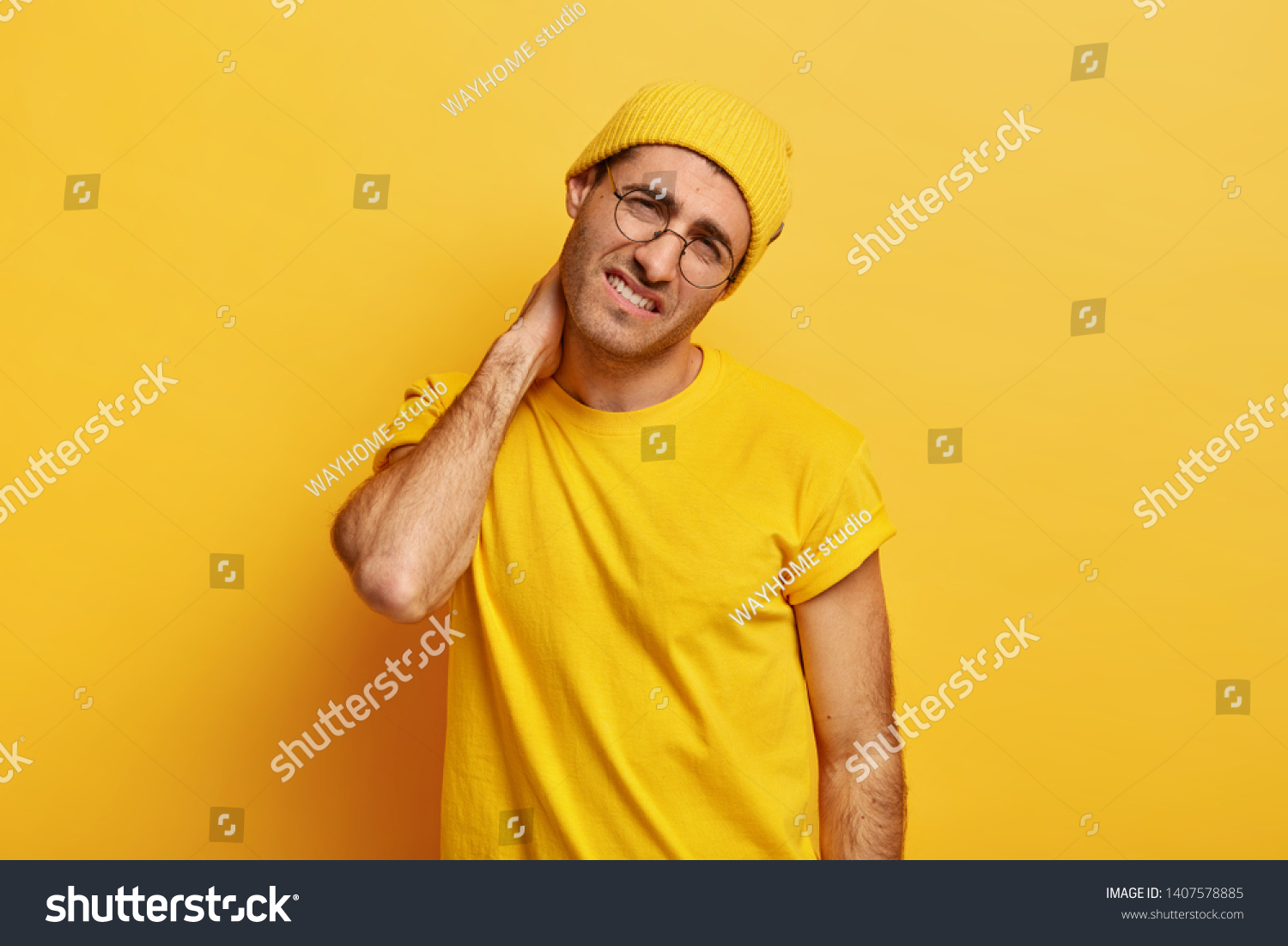 Overworked dissatisfied young European man keeps hand on neck, feels spasm in neck, tilts head, wears yellow hat and t shirt, poses over yellow background. People, tiredness and problem concept #1407578885
