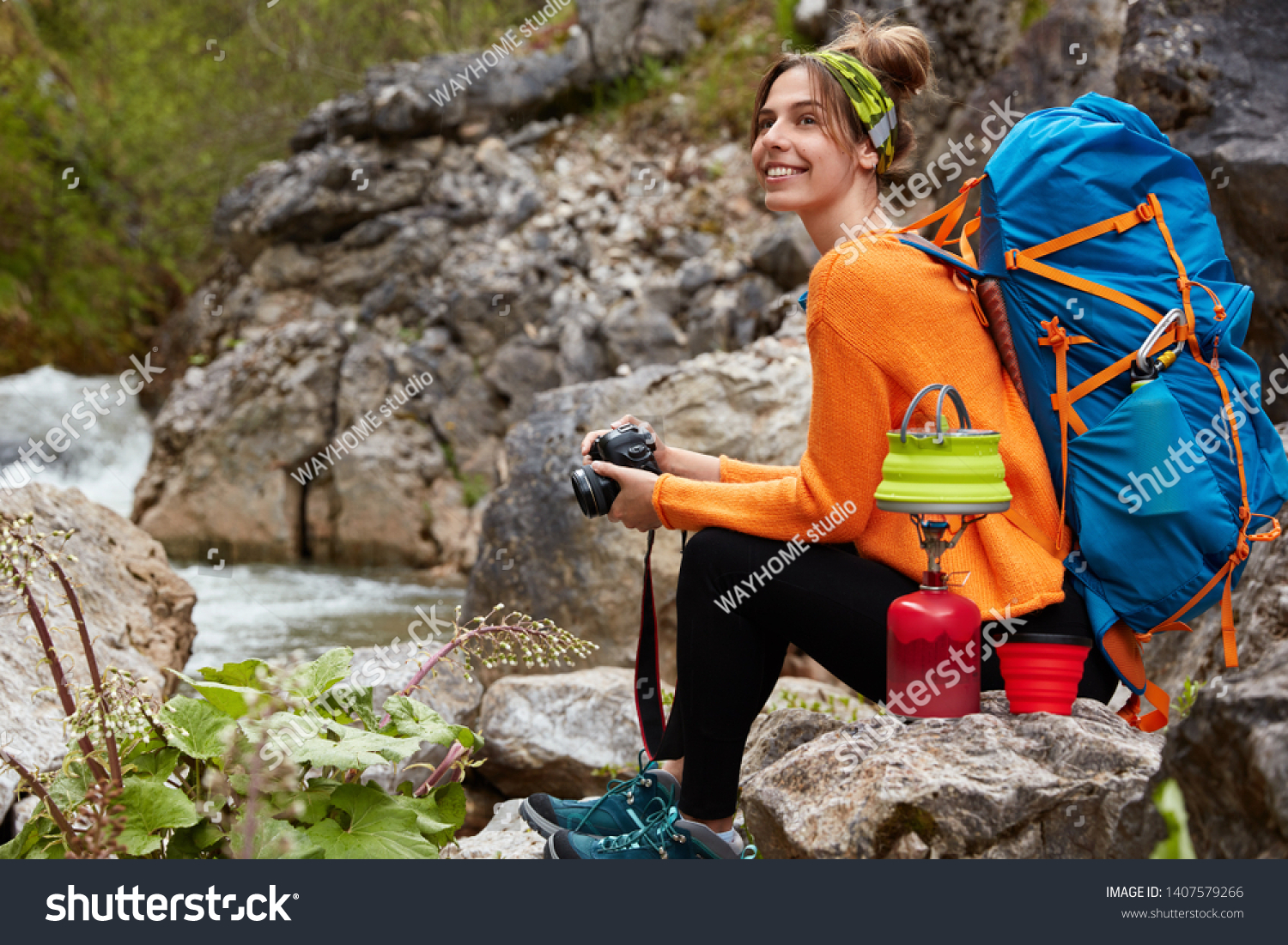 Horizontal view of cheerful pleased woman sits near rock pool, holds modern camera, prepares hot drink, enjoys camping and traveling, wears active wear, has rucksack on back. Rest, lifestyle, trekking #1407579266