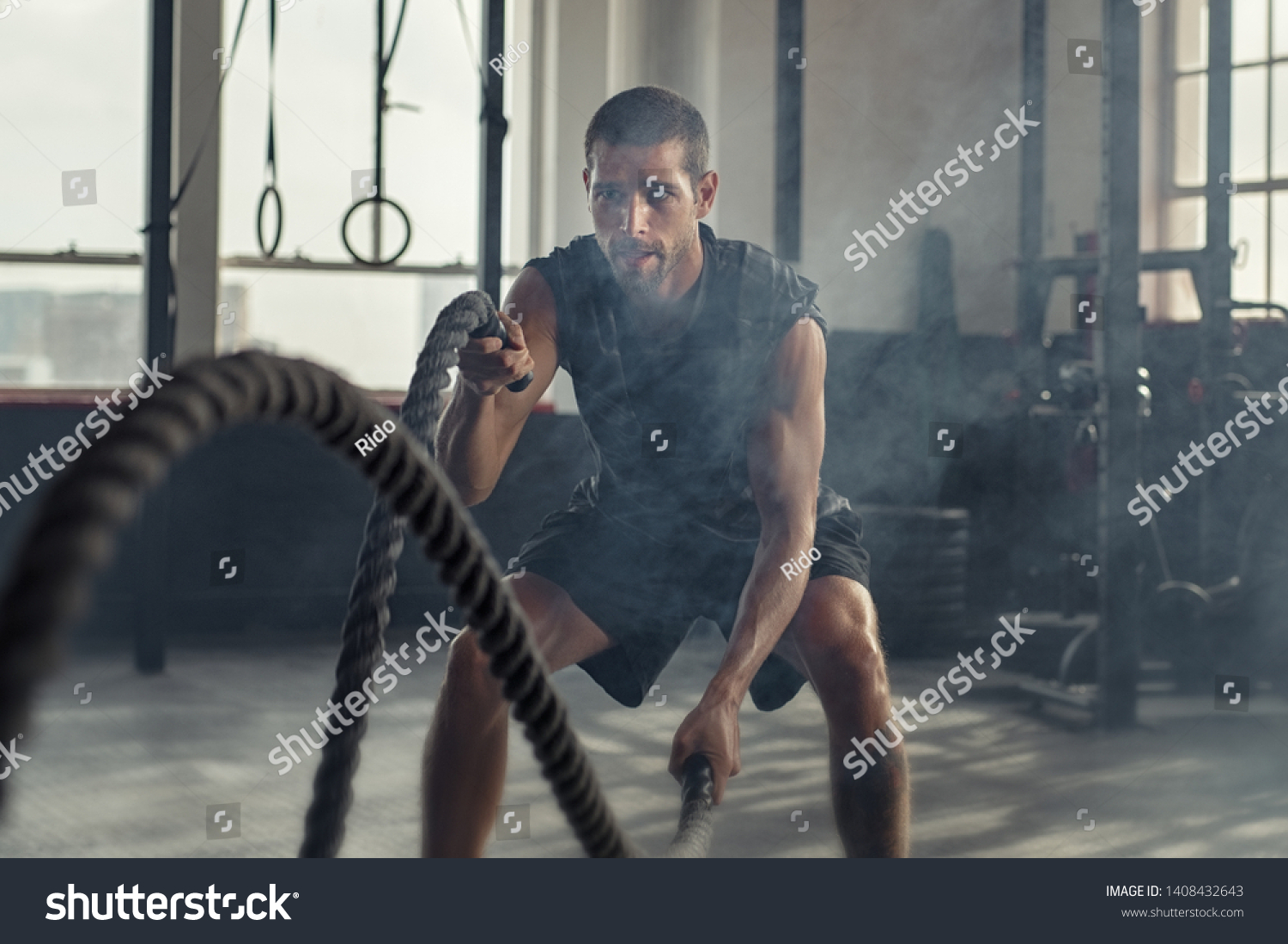 Strong young man working out with battle ropes in a crossfit gym. Muscular sportsman doing cross excursion with ropes in workout gym. Determined guy using battle rope while doing physical training. #1408432643