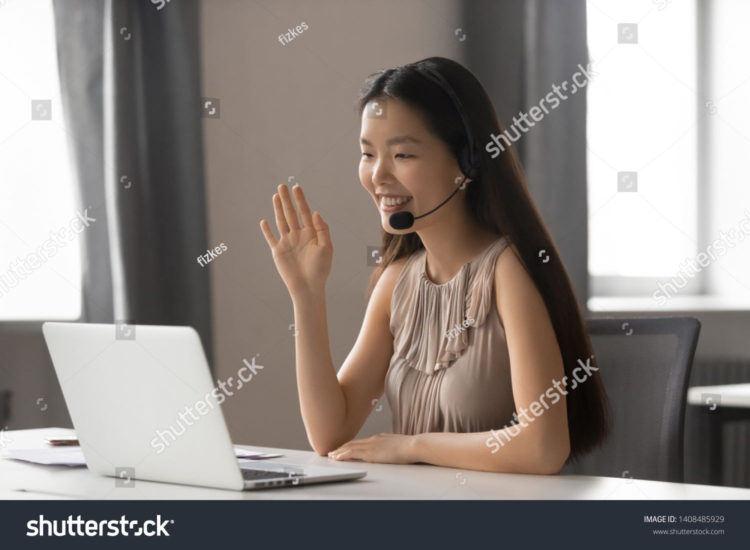 Smiling asian businesswoman wearing wireless headset waving hand doing video chat looking at laptop consulting customer sitting at workplace, female call center service support agent talking  #1408485929