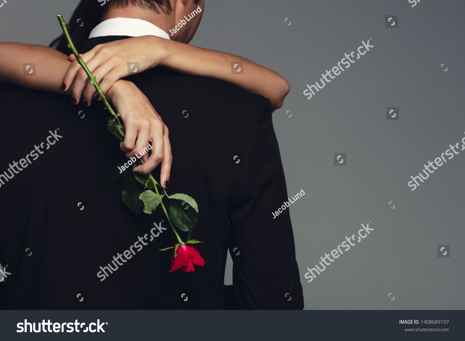 Rear view of woman holding a rose embracing man in black suit. Loving couple embracing on grey background. #1408689197