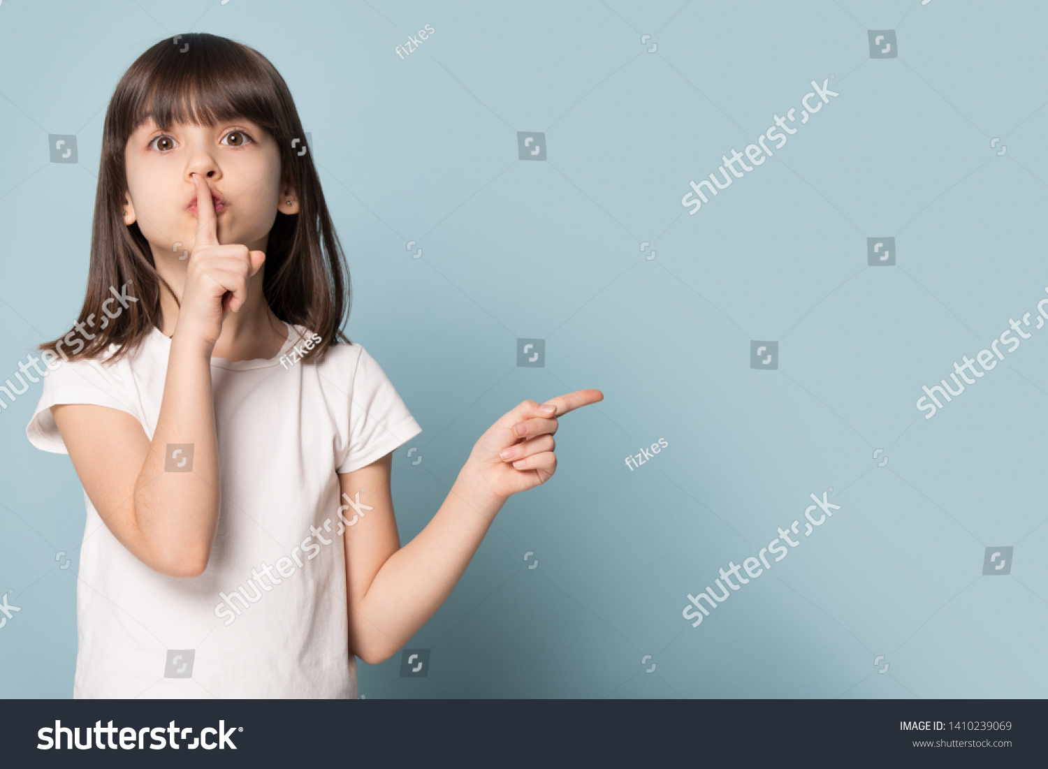 Adorable six years old little girl holding finger on lips symbol of hush gesture of asking to be quiet. Silence or secret concept image isolated on blue studio background with copy free space for text #1410239069