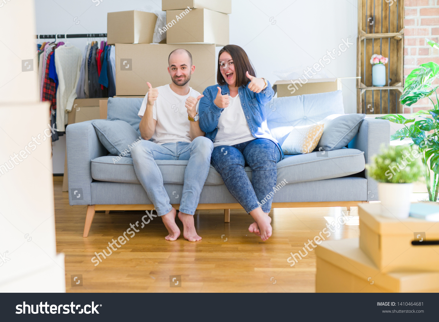 Young couple sitting on the sofa arround cardboard boxes moving to a new house success sign doing positive gesture with hand, thumbs up smiling and happy. Cheerful expression and winner gesture. #1410464681