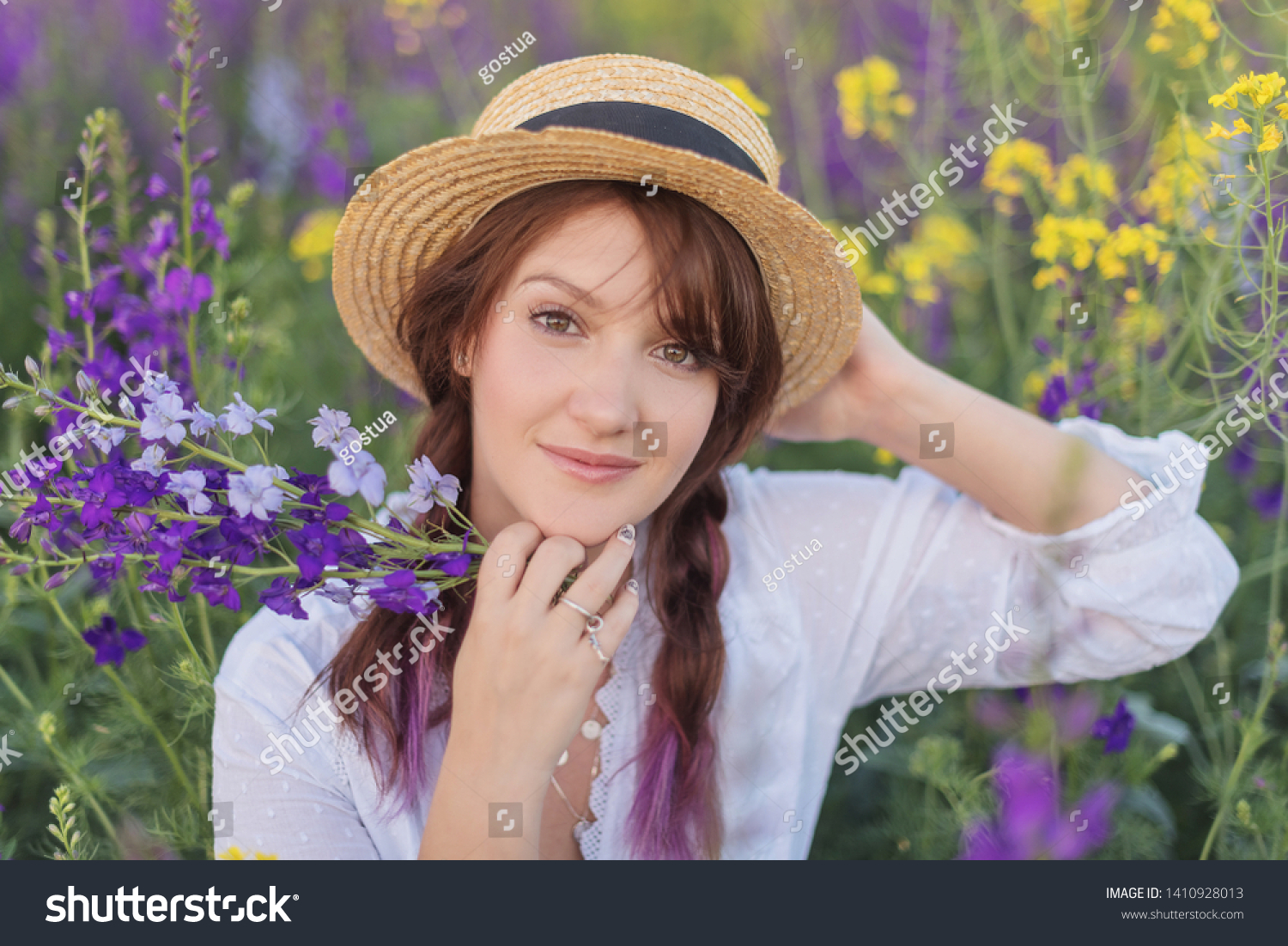 Portrait of a beautiful brunette girl with long hair and pigtails in a straw hat and white dress that holding wild flower in a blossoming field
 #1410928013