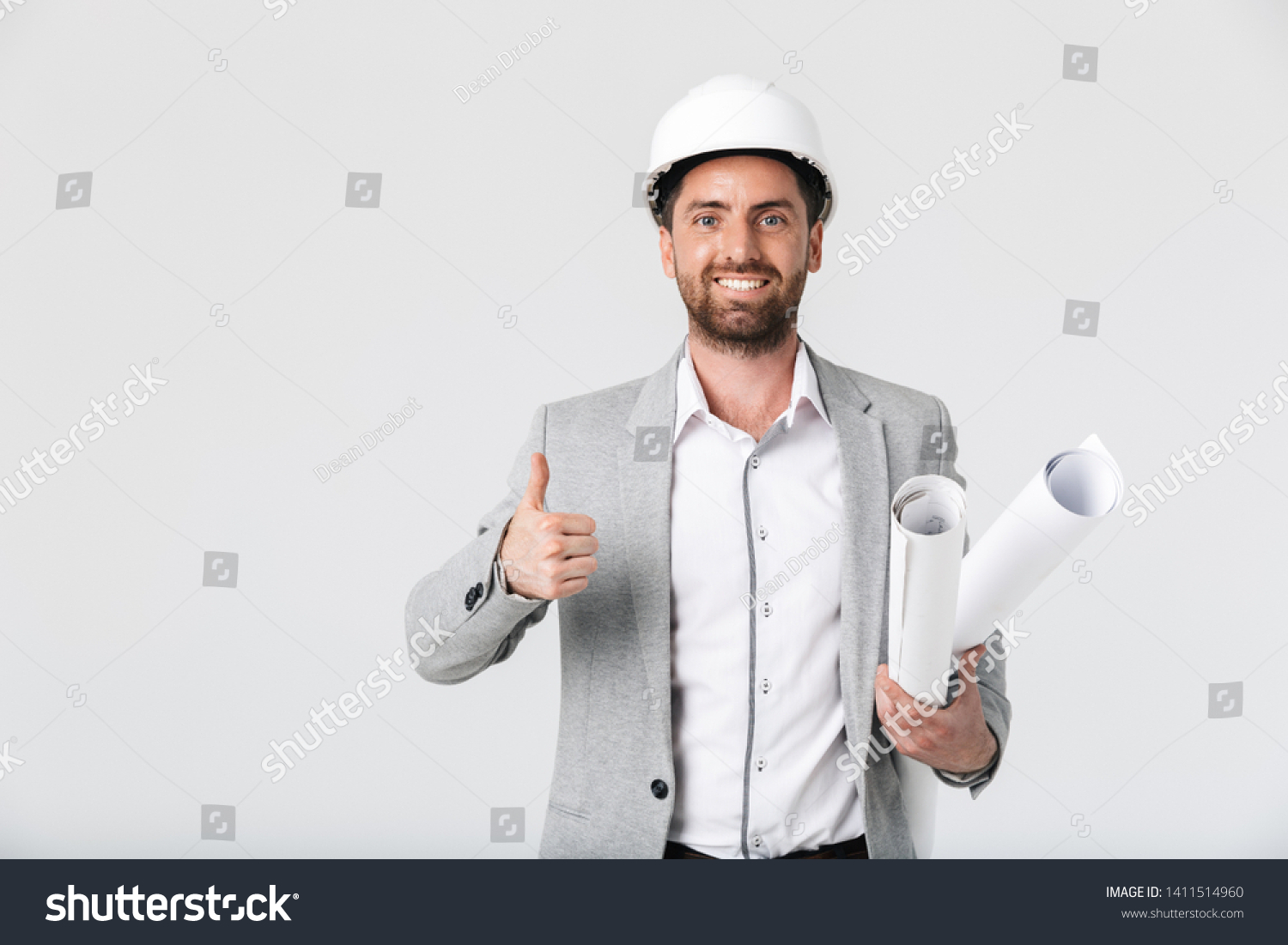 Confident bearded man builder wearing suit and hardhat standing isolated over white background, carrying blueprints, thumbs up #1411514960