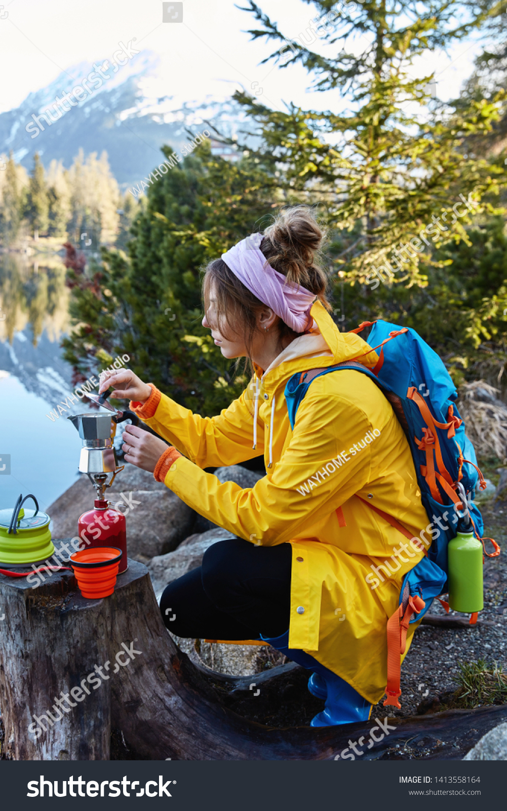 Restful female traveler makes coffee on camping stove, poses near stump, has break after wandering, poses against wonderful nature with lake, mountains and fir trees, enjoys life and traveling #1413558164