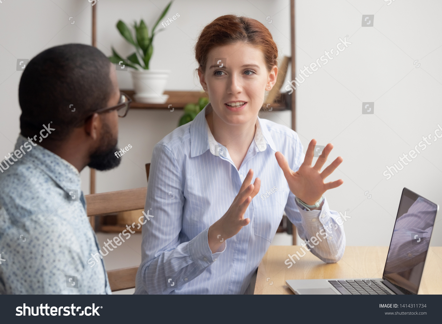 Two diverse businesspeople chatting sitting behind laptop in office. Excited caucasian female sharing ideas or startup business plan with black male coworker. Informal conversation, work break concept