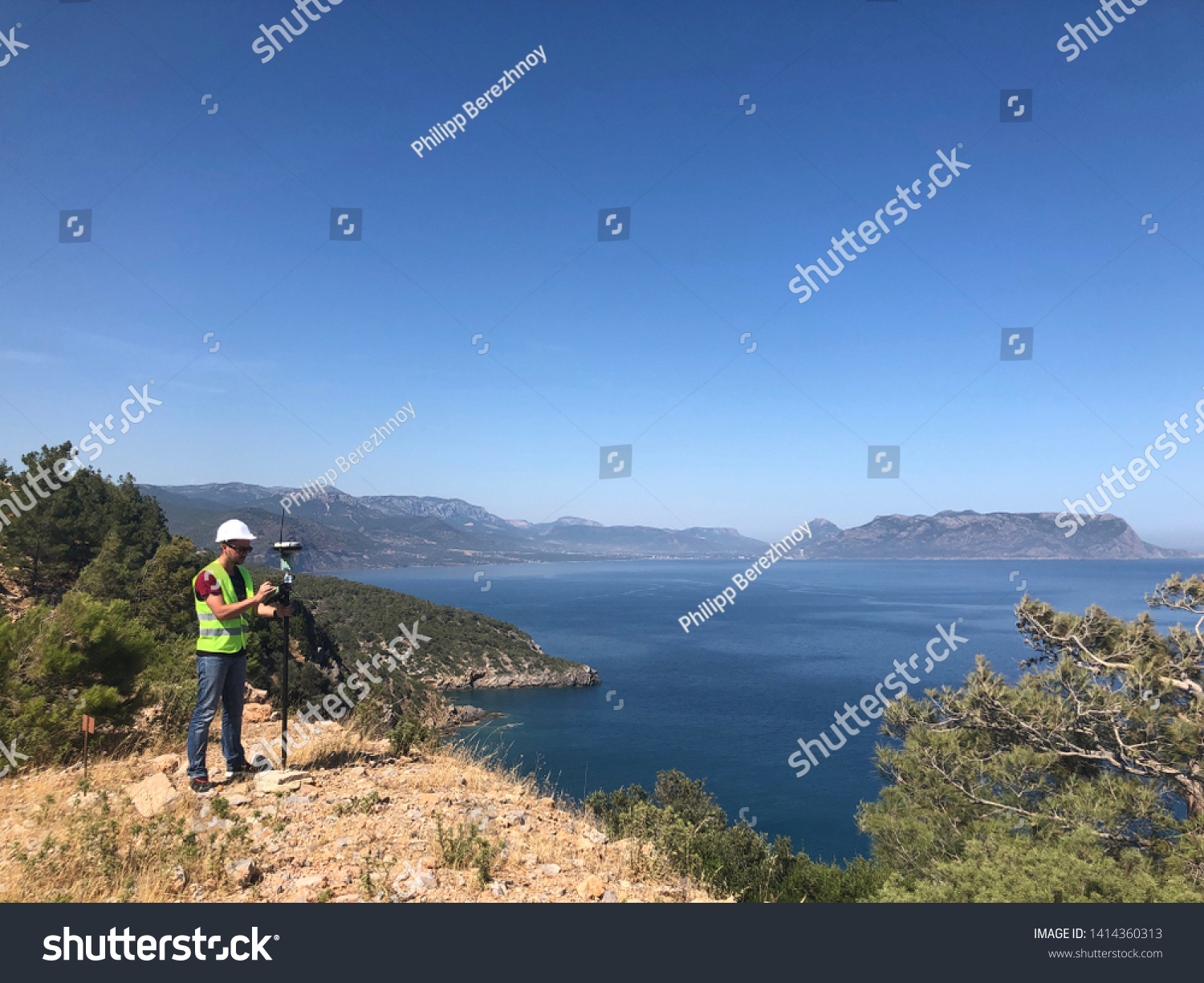 Geodetic engineer surveyor in white hard hat doing measurements with GNSS satellite receiver at the hill top above the sea #1414360313