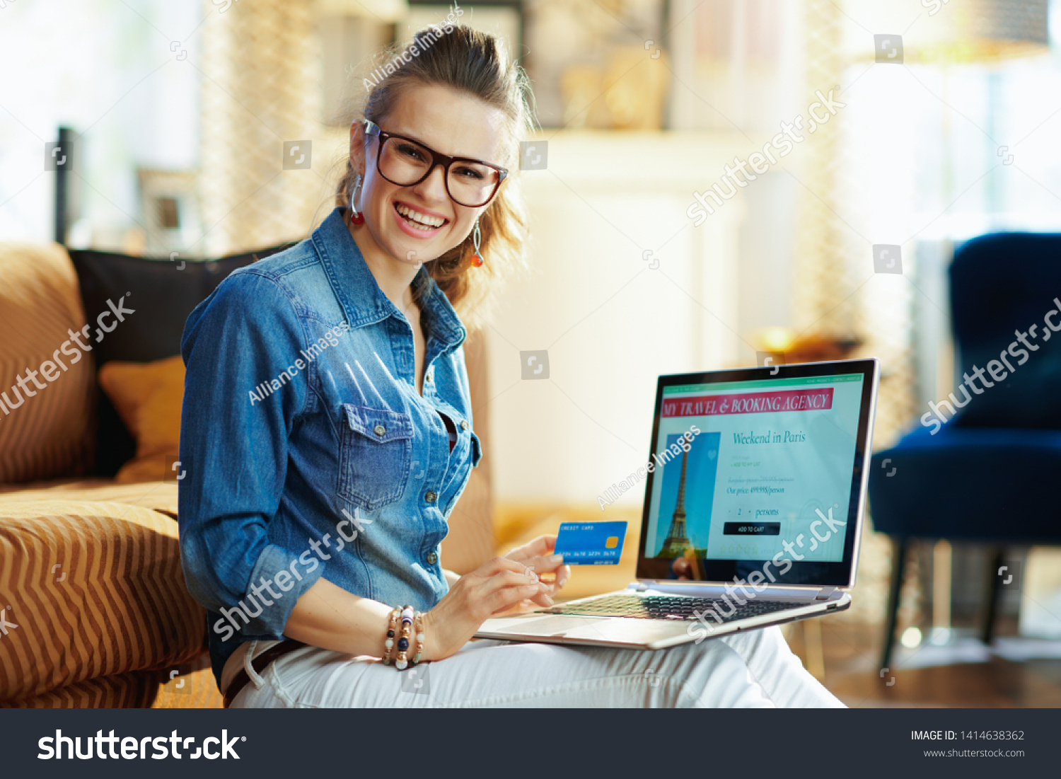 happy elegant woman with opened on laptop online travel & booking agency site holding credit card in the modern living room in sunny summer day. A fake website is made for illustrative purposes. #1414638362