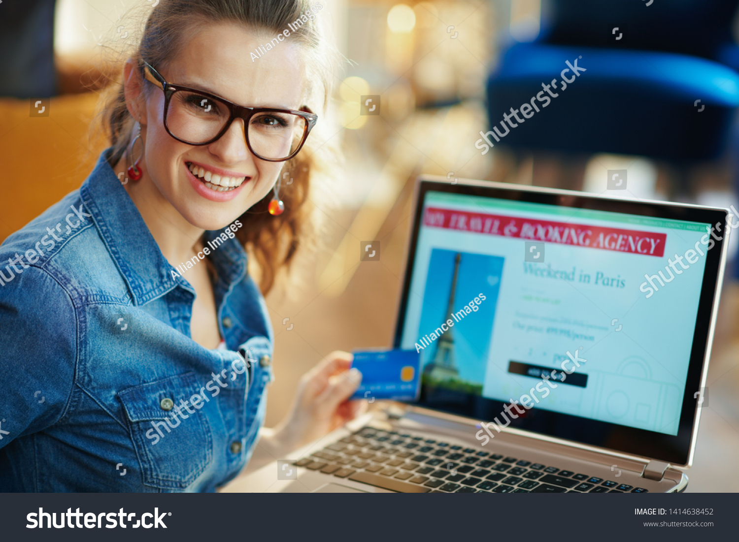smiling modern woman with opened on laptop online travel & booking agency site holding blue credit card in the modern living room in sunny summer day. A fake website is made for illustrative purposes #1414638452