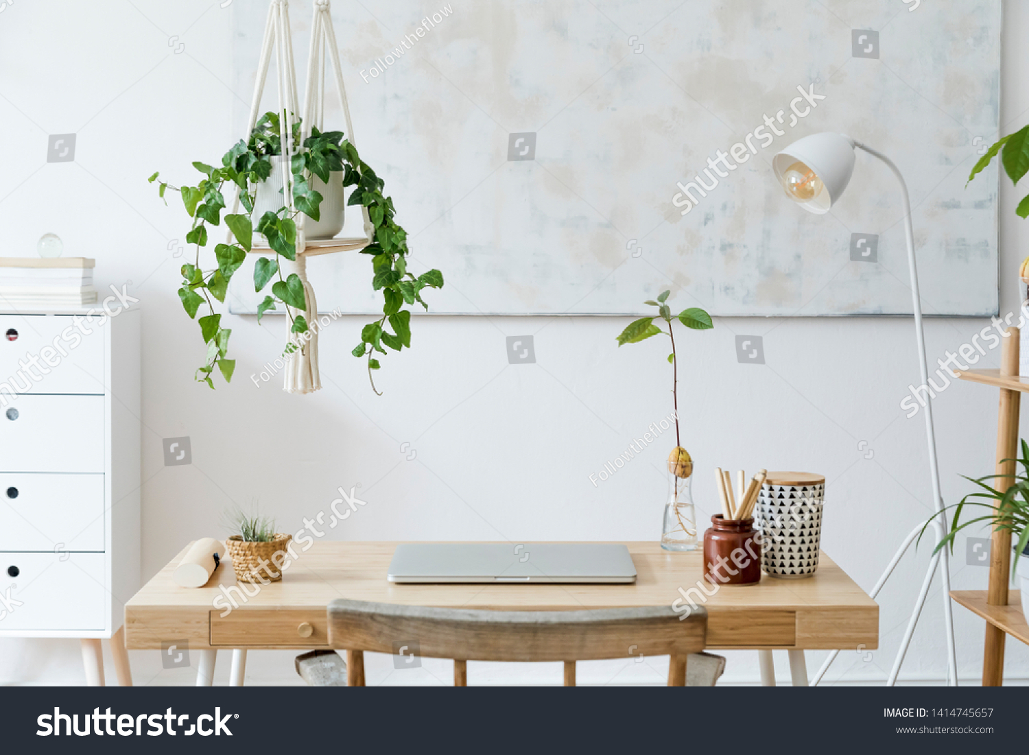 Stylish and boho home interior of open work space with wooden desk, chair, lamp, laptop and white shelf. Design and elegant personal accessories. Botany and minimalistic home decor with plants. #1414745657
