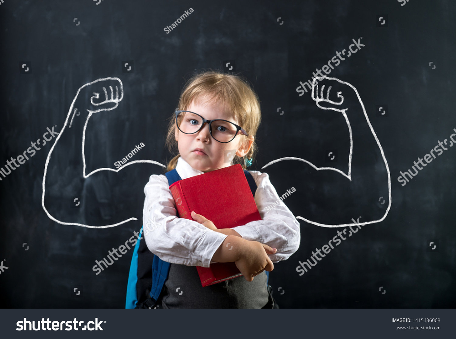 Cute child girl in school uniform and glasses. Go to school for the first time. Child with school bag and book. Kid in class room near chalkboard with muscles on it. Back to school #1415436068