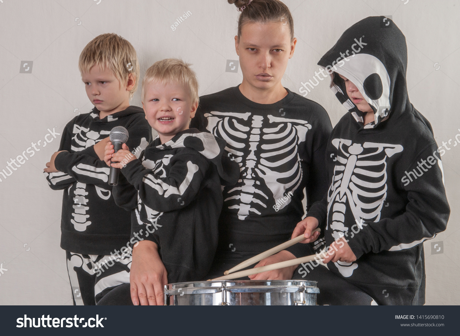 Happy friendly family of musicians in carnival costumes, boys and young mother play drum and try to sing with microphone. Black suit with image of skeletons. Classic halloween costume. Funny children #1415690810