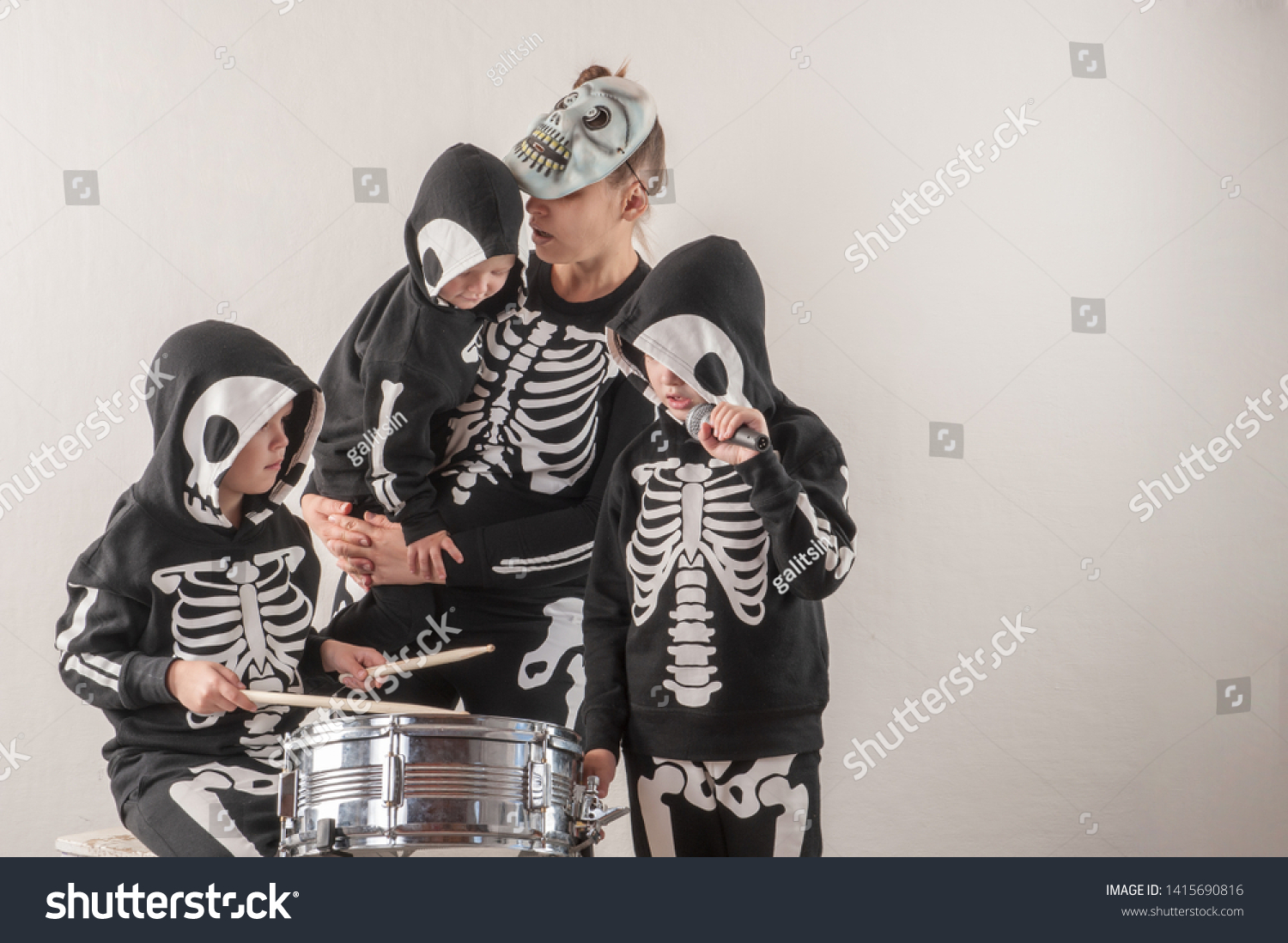 Happy friendly family of musicians in carnival costumes, boys and young mother play drum and try to sing with microphone. Black suit with image of skeletons. Classic halloween costume. Funny children #1415690816