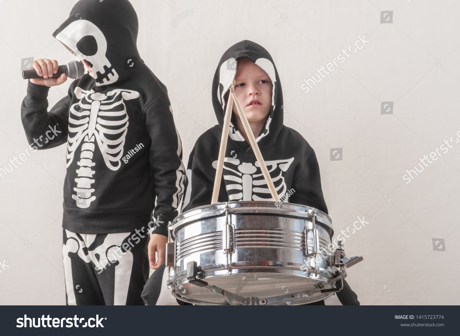 Happy friendly family of musicians in carnival costumes, boys play drum and try to sing with microphone. Black suit with image of skeletons. Classic halloween costume. Funny children #1415723774