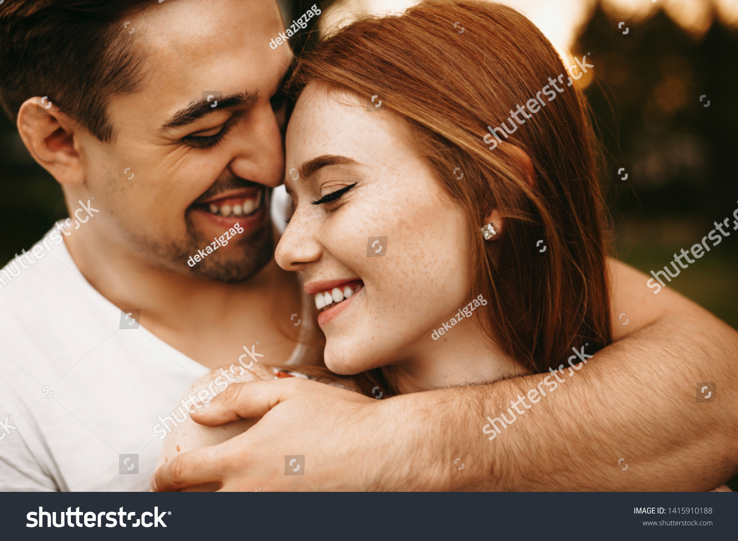 Close up portrait of a amazing young female with red hair and freckles smiling with closed eyes while being embraced from back by her boyfriend closely outside. #1415910188