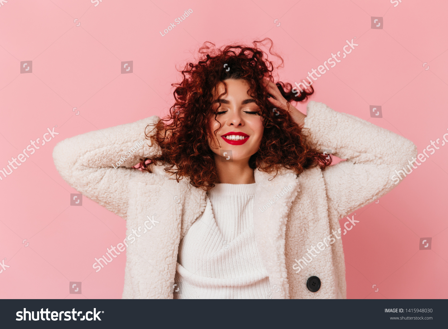 Portrait of charming lady with red lipstick and snow-white smile dressed in bright eco-coat and touching her curly hair on pink background #1415948030