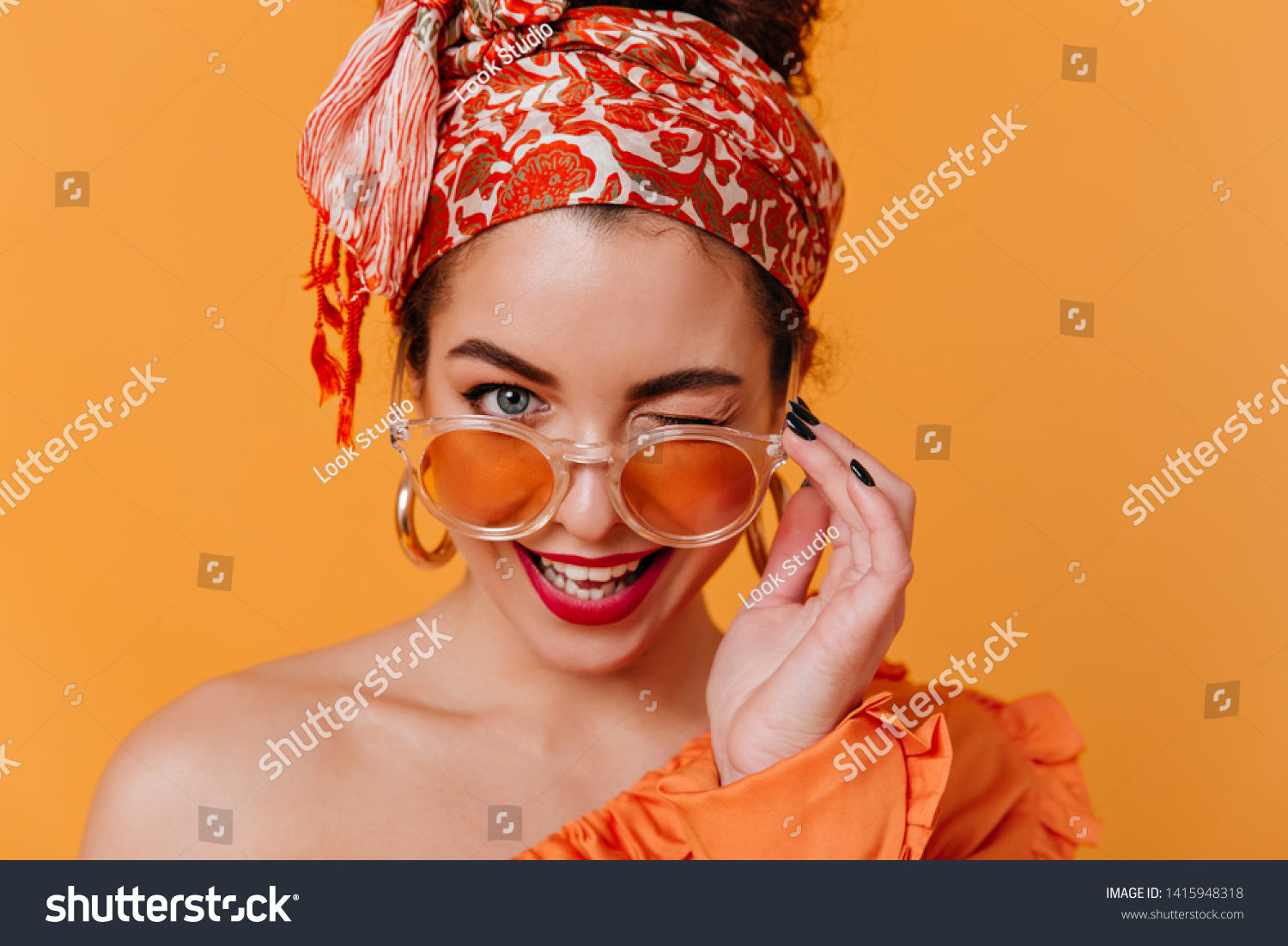 Lovely woman in African-style massive earrings and headband removes her orange glasses and winks coquettishly #1415948318
