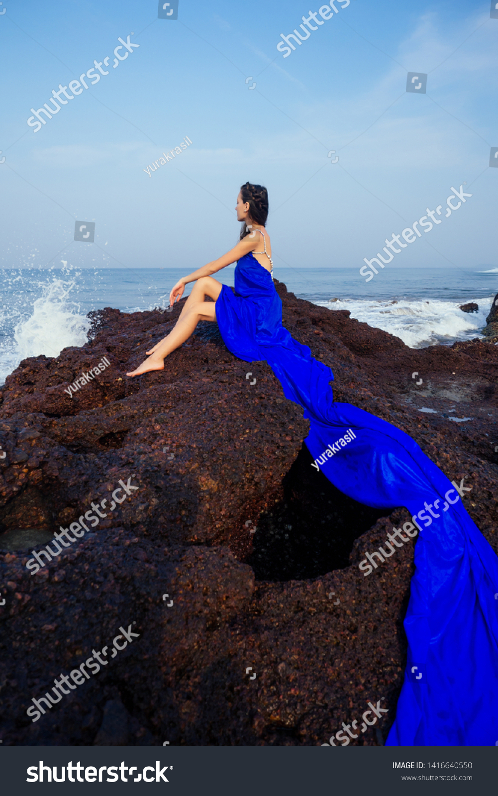 fashion pretty female model posing on a beach with rocks in a long butterfly chameleon dress waterfall of skirt plume train.sensual perfume with a beautiful and young brunette woman wave tropical sand #1416640550
