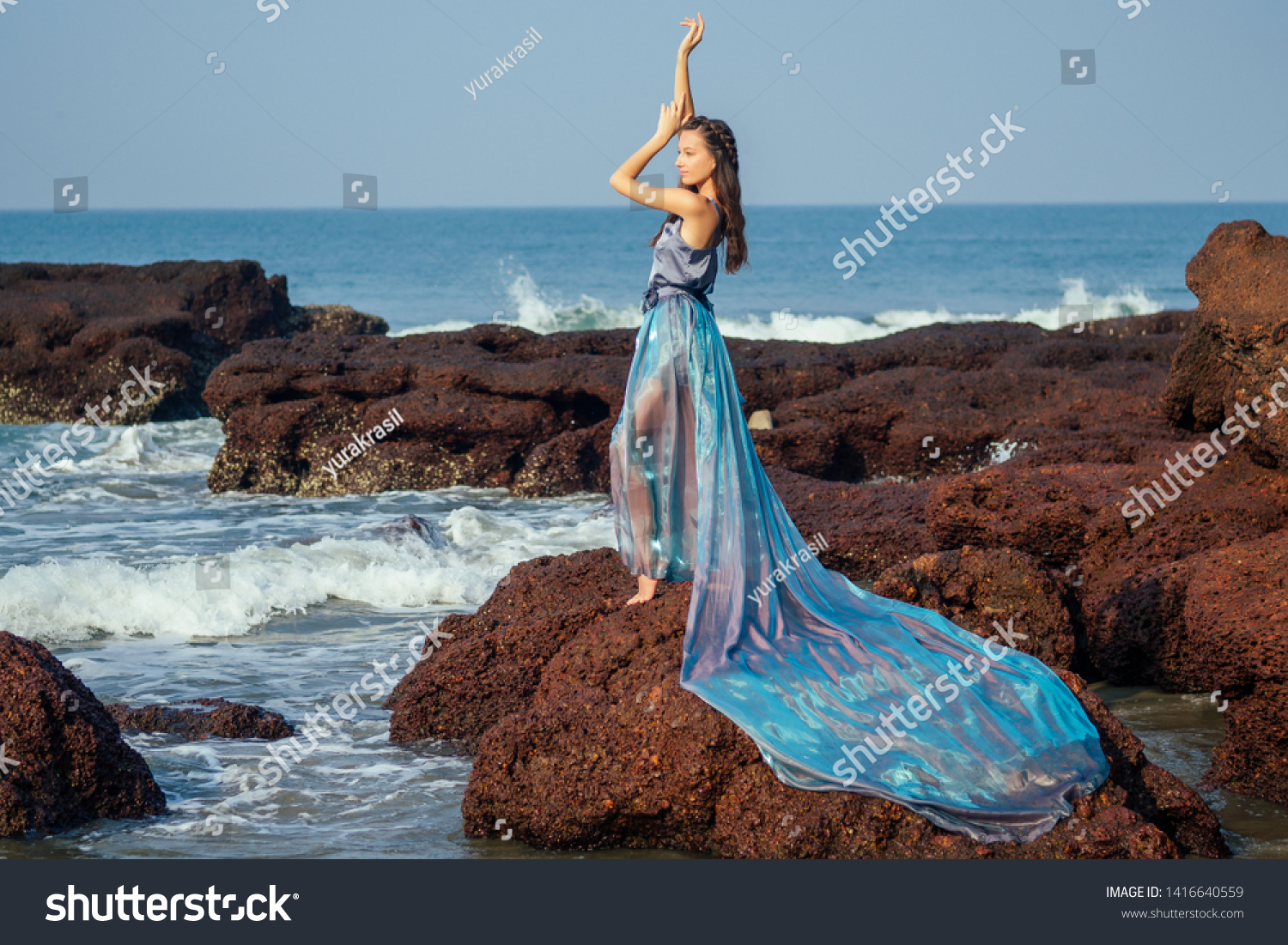 fashion pretty female model posing on a beach with rocks in a long butterfly chameleon dress waterfall of skirt plume train.sensual perfume with a beautiful and young brunette woman wave tropical sand #1416640559