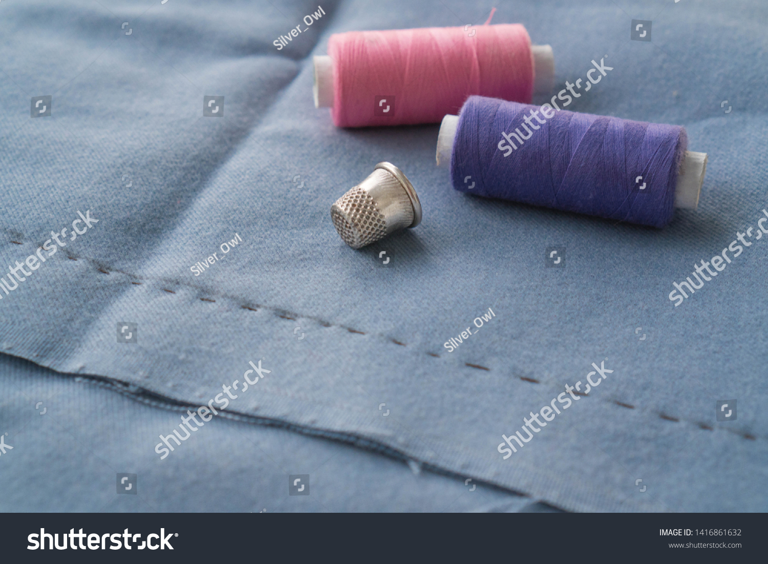 Cut part of skirt with a sewn tuck, thimble and two spools of thread. Two spools of pink and purple threads and tailor tool are lying on the blue basting fabric #1416861632
