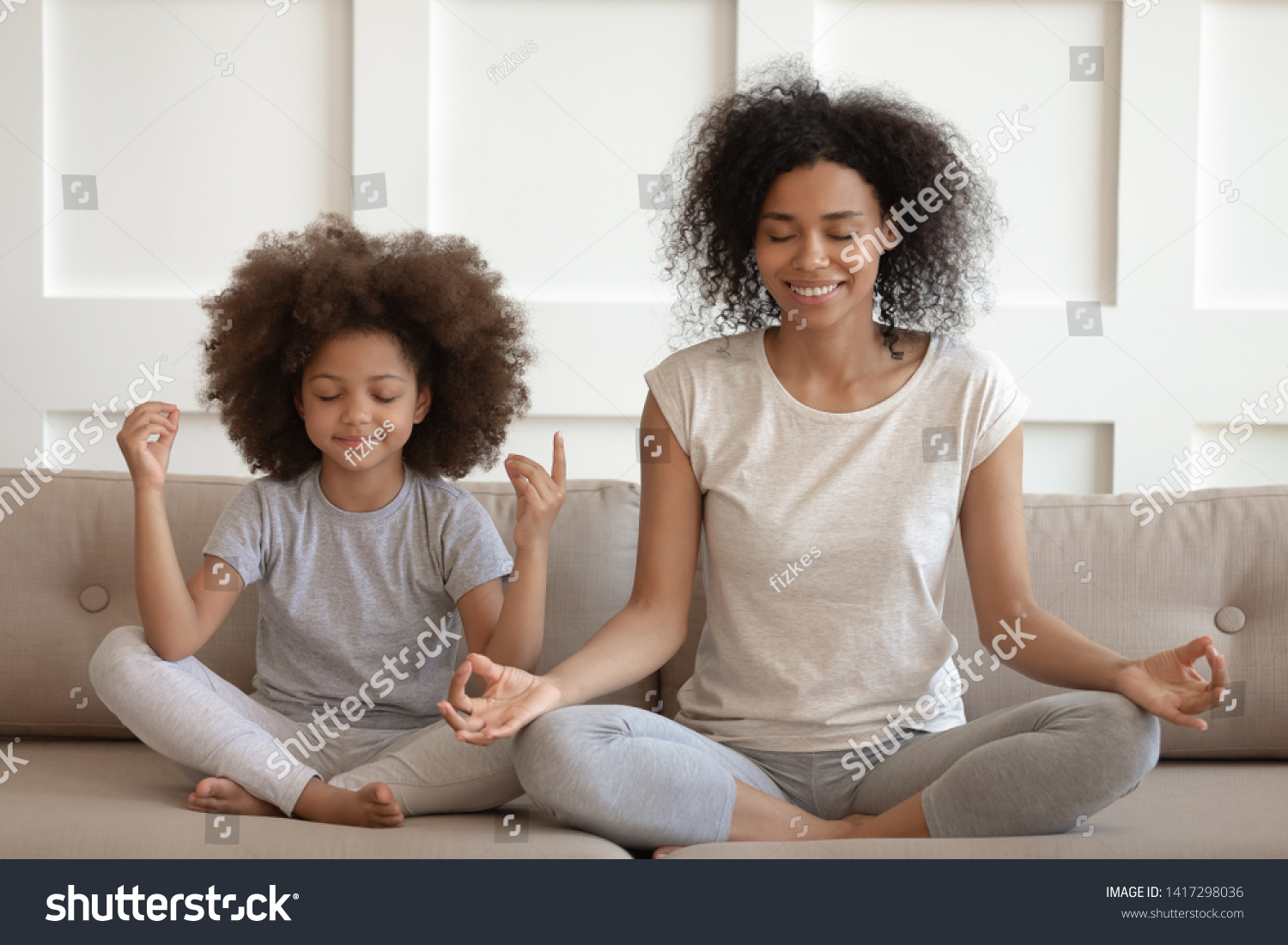 Healthy calm african american mother teaching meditation sit with cute small child daughter on sofa, mindful happy black mom and kid girl doing yoga exercises relaxing together in lotus pose at home #1417298036