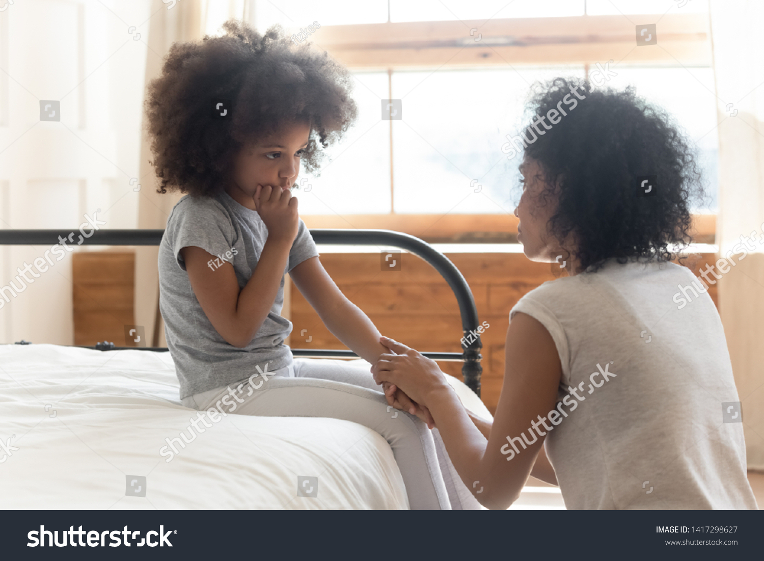 Caring worried african american mother holding hand of sad little mixed race daughter talking giving support and comfort, black mom foster parent consoling small kid being bullied sit on bed at home #1417298627